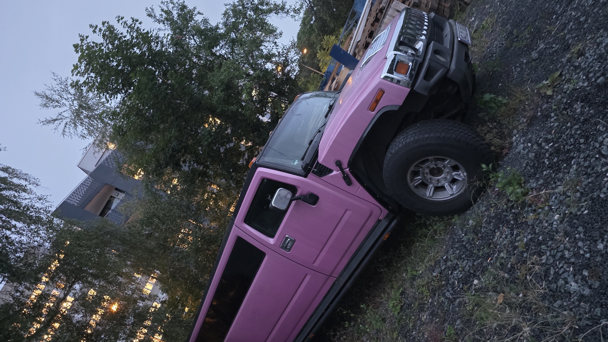 A bright pink Hummer limousine is parked on a gravel lot next to a building and trees. The building has many windows with lights on inside. The sky is a pale blue, indicating either early morning or dusk. The limousine is angled in the frame, with its front right wheel closest to the viewer. The ground is covered in small, dark gray gravel and some sparse green vegetation. Pallets are stacked in the background to the right of the limousine. The scene appears to be in an urban or suburban area, given the presence of the building and the limousine.
