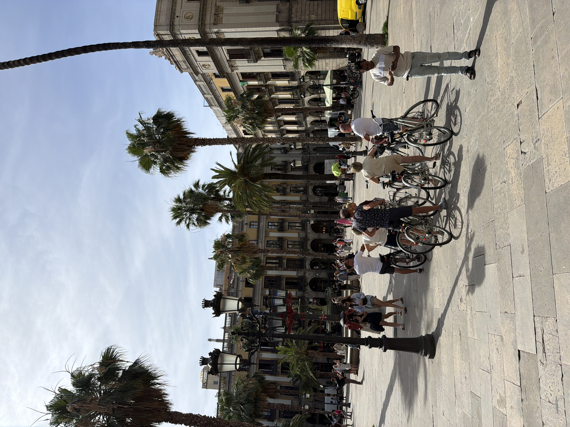 This image captures a sunny day in Plaça Catalunya, Barcelona, Spain. The foreground features a cobblestone plaza with several people on bicycles, some standing and others preparing to ride. A man in a light-colored shirt and jeans stands to the right, looking at his phone. In the mid-ground, a large ornate lamppost stands prominently. Behind it, a fountain with red accents is visible, surrounded by more people and palm trees. The background is dominated by grand, multi-story buildings with arched windows and balconies, characteristic of the city's architecture. A bright yellow van is parked on the right side of the image, near the buildings. Lush palm trees are scattered throughout the scene, indicating a warm climate. The sky is mostly clear with some scattered clouds.