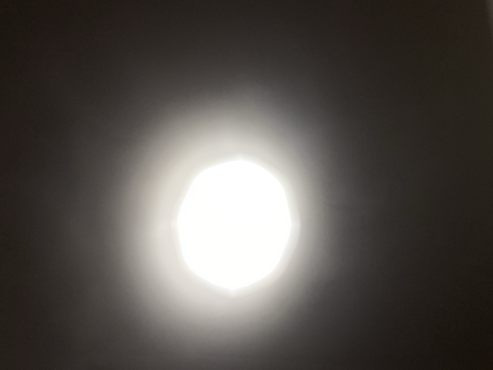 The image displays a bright, circular light source emanating from the center against a dark background. The light source is not a perfect circle, but rather has an irregular, polygonal shape with several discernible facets, suggesting it might be a lamp or a light fixture with a patterned cover or a natural light source with an obstruction. The light is intensely bright, creating a halo effect and significant glare that washes out details in the immediate vicinity.
The background is a gradient of dark tones, appearing almost black at the edges and subtly lightening as it approaches the central light source. This suggests the image was taken in a dimly lit environment where the bright light is the dominant element. There are no discernible objects, people, or specific scene elements other than the light itself and the surrounding darkness.
The lighting implies that the photo was taken indoors, possibly at night or in a room with controlled lighting, as there are no environmental cues like sunlight or shadows that would indicate a specific time of day or outdoor setting. The overall mood conveyed by the image is one of intense illumination, possibly an abstract representation of light or a close-up shot of a light source. No text is visible in the image.
