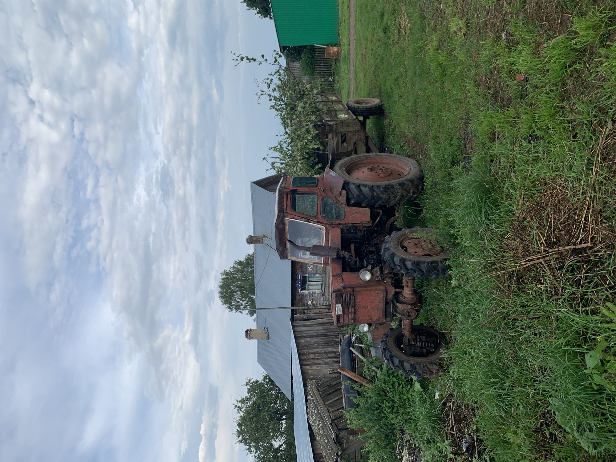 An aged, red tractor with a wooden trailer filled with branches is parked on a grassy area in front of a rustic wooden building. The trailer and tractor show signs of wear and tear, with rust and dirt visible on their surfaces. Behind the tractor, a weathered wooden structure with a grey metal roof and two chimneys stands under a cloudy sky. To the right of the tractor, a bright green metal building is visible. The surrounding environment is rural, with overgrown grass and vegetation. The overall scene depicts a rural setting in the village of Panteleevka, Russia, evoking a sense of agricultural life and perhaps neglect or disuse of the equipment.