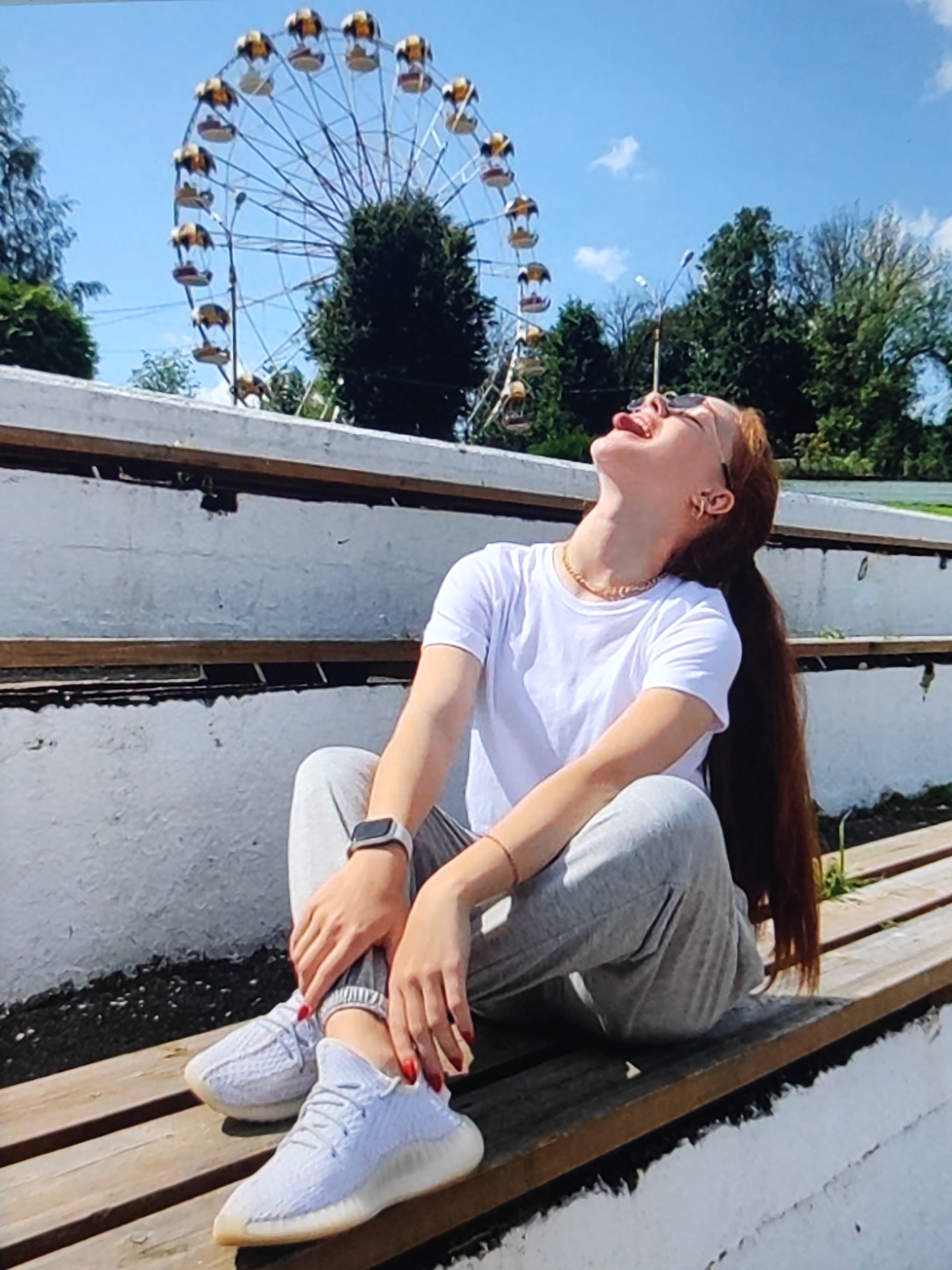A young woman with long, auburn hair is sitting on a wooden bench outdoors, looking upwards with her mouth open in a laugh or shout. She is wearing a white t-shirt, grey sweatpants, and white sneakers. A smartwatch is visible on her left wrist. Behind her, a Ferris wheel is visible against a bright blue sky with scattered clouds. Trees and foliage are also present in the background. The bench appears to be part of a tiered seating area, possibly in a park or recreational area.
