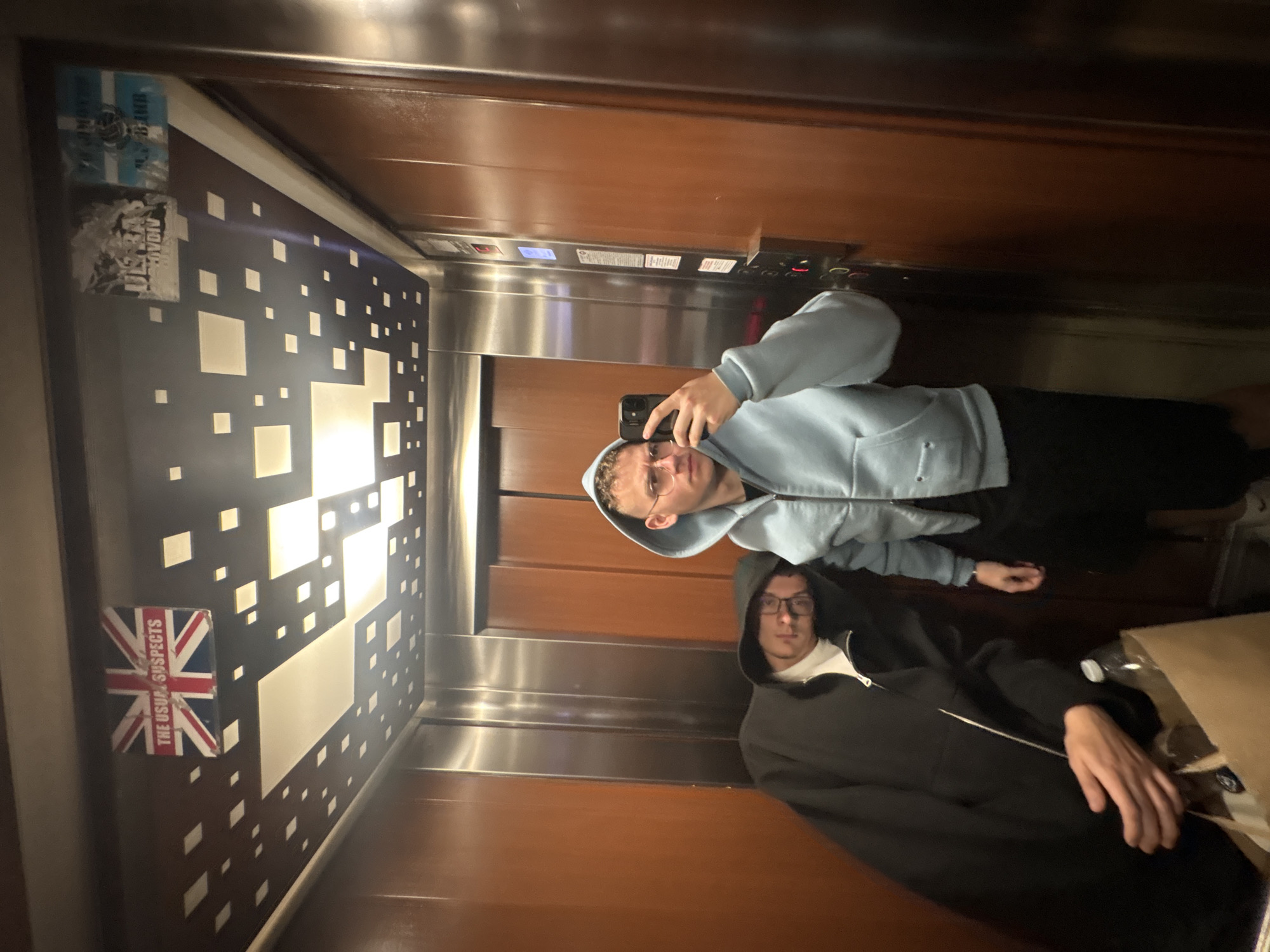 The media file shows a mirror selfie taken by two young men inside an elevator. The image is rotated counter-clockwise.
**Subjects:** Two young men are present. The one taking the photo, reflected in the mirror, wears a light blue hooded sweatshirt and glasses, holding a black smartphone. The second man stands behind him, wearing a dark (likely black) hooded sweatshirt with the hood up, and also wears glasses.
**Scene/Setting:** The setting is an elevator. Its interior features a decorative back wall with a pattern of varying white squares on a dark background, some of which are illuminated. The side walls and ceiling are paneled with wood or a similar material, and there are polished metallic surfaces, particularly around the elevator doors and control panel.
**Activity or event taking place:** The two men are posing for a casual mirror selfie within the elevator.
**Notable details:** The lighting is artificial, suggesting an indoor setting without natural light. On what appears to be the left wall of the elevator (in corrected orientation), there are several stickers. One prominent sticker is a Union Jack flag. Above it are partially torn blue and white stickers. A brown paper bag and a plastic water bottle are visible on the floor near the second man. The elevator control panel, with its buttons and floor indicators, is visible on the right. The location context of Plovdiv, Bulgaria, is not explicitly visible within the image itself.
**Visible text:** The Union Jack sticker clearly displays the text "THE USUAL SUSPECTS". Other stickers contain unreadable or partially obscured text. The elevator control panel features unreadable labels on its buttons and indicators.