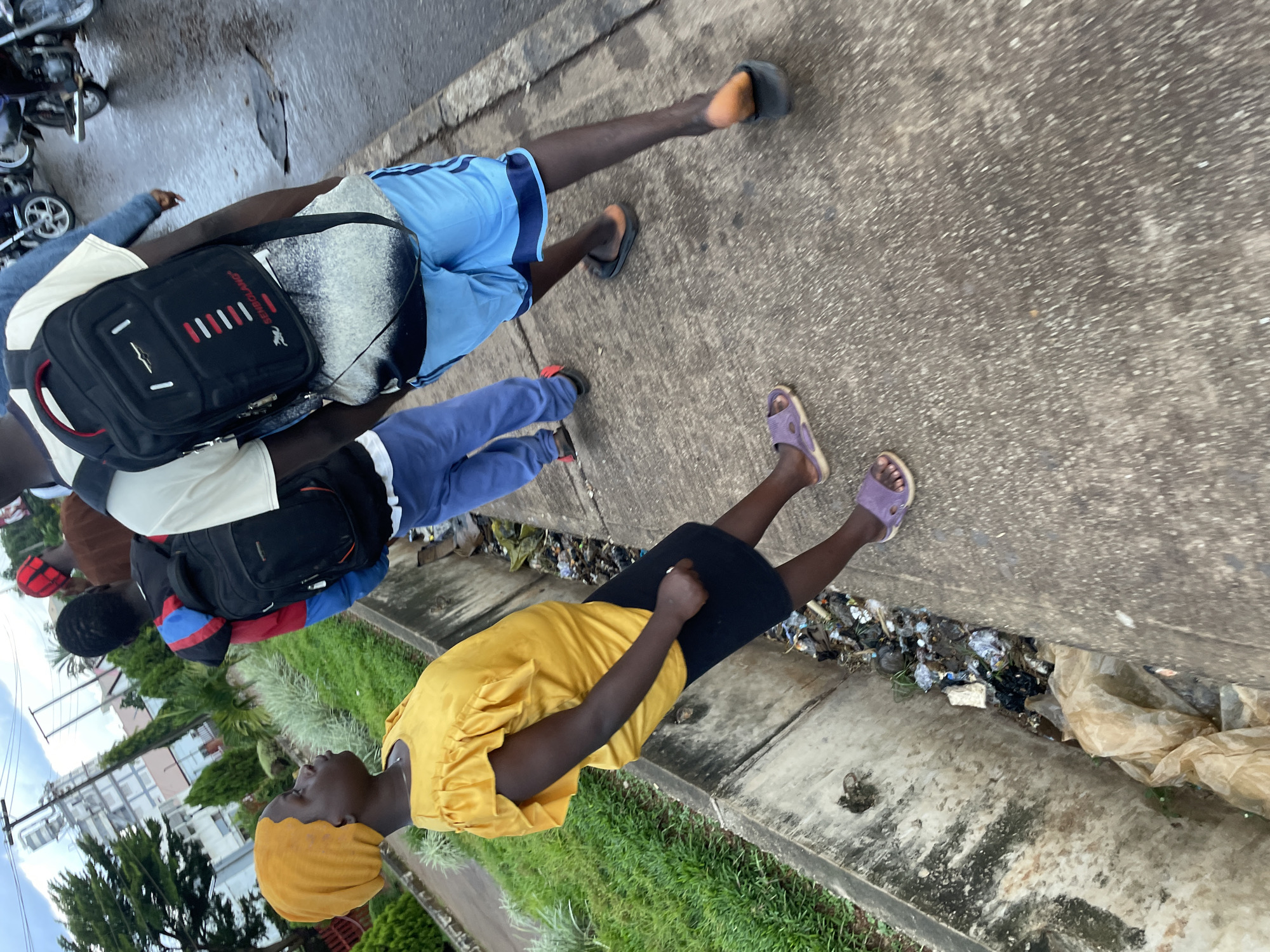 The image depicts a street scene in Barakin Lerawi, Nigeria, on an overcast day. In the foreground, a young girl wearing a yellow top and a black skirt walks along a concrete sidewalk. To her right, a portion of a drainage ditch filled with debris is visible. Behind her, several individuals are walking, one carrying a black backpack. They appear to be moving in the same direction. The background shows a street with some vegetation and buildings, including what appears to be an industrial structure with tall pipes. Motorcycles are also visible in the background, suggesting an urban environment. The overall atmosphere of the image is one of everyday life on a street in a Nigerian city.