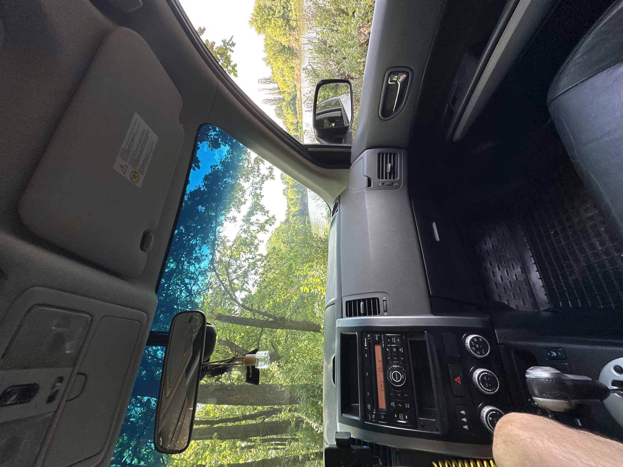 The image shows the interior of a car, looking out through the windshield. The car is parked in a natural setting, with a body of water and lush green trees visible outside. The dashboard of the car is visible in the foreground, with the radio and climate controls showing. A person's arm is visible on the left side of the image, resting near the gear shift. The overall impression is of a peaceful, scenic location. The location is Shatava, Ukraine.