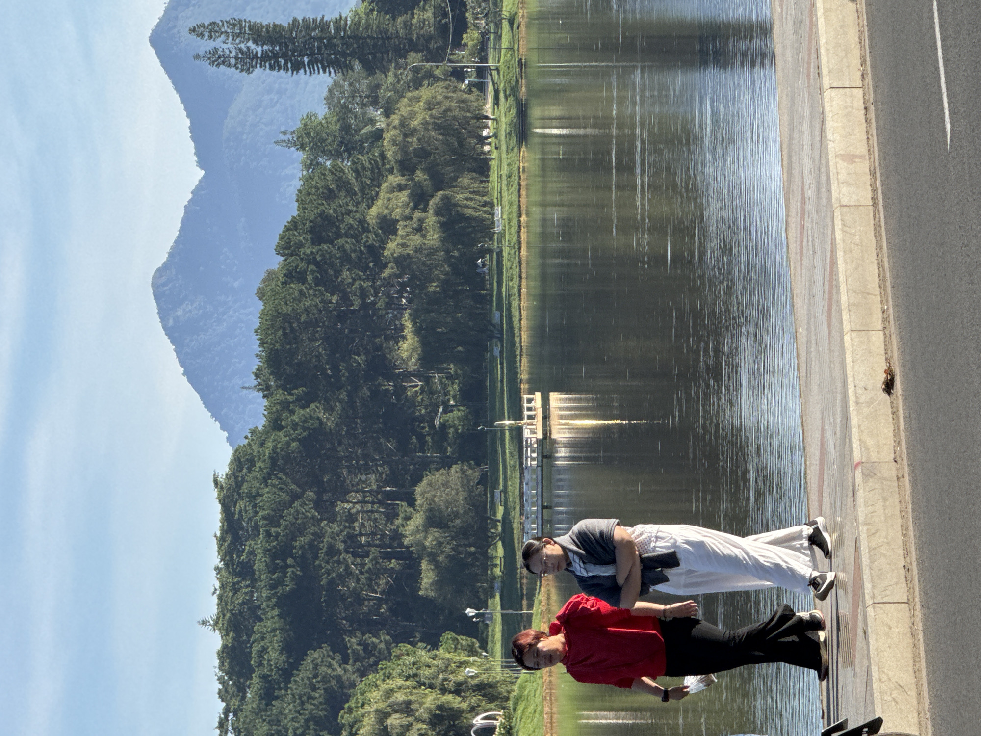 Two women are walking on a paved pathway next to a calm lake. The woman on the left wears a red hooded sweatshirt and black pants. The woman on the right wears a grey shawl over a white outfit and black sneakers. Behind them, the lake reflects trees and mountains. The environment appears to be a park in Da Lat, Vietnam, known for its lakes and pine forests. The sun is shining, casting a warm light on the scene. The pathway has a concrete curb and a textured surface. The background shows a dense forest of pine trees and distant blue mountains under a clear sky. A small wooden pier is visible in the lake.