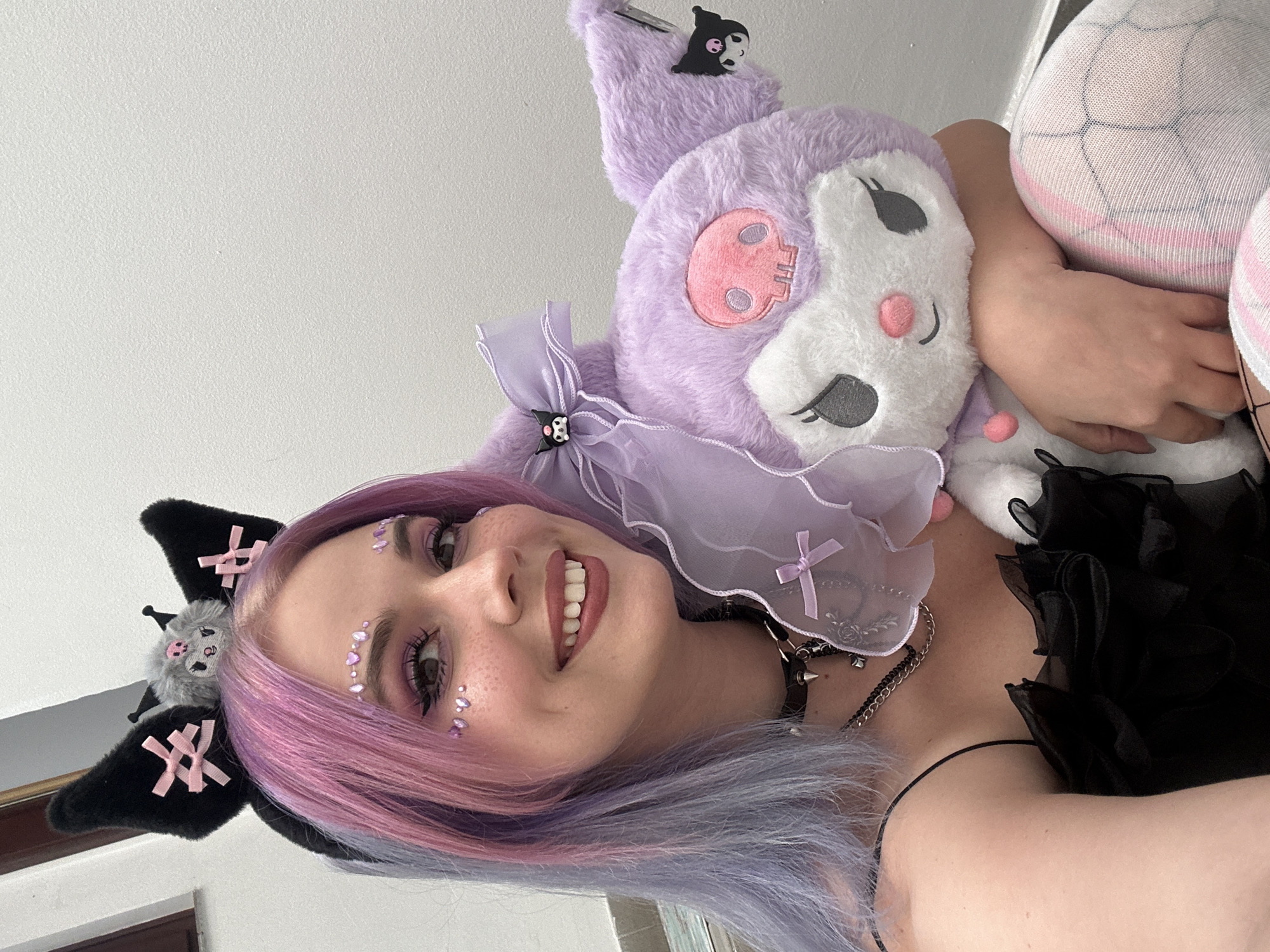A young woman is holding a stuffed Kuromi character toy while taking a selfie in an indoor location. The woman has dyed pink and blue hair and is wearing black cat-ear hair accessories, as well as decorative face jewels. She has visible freckles on her face. The woman is also wearing layered necklaces and a black top. The stuffed Kuromi is light purple with a white face, adorned with a pink skull design and a layered purple bow. The scene is not discernible as City of Zagreb, Croatia.