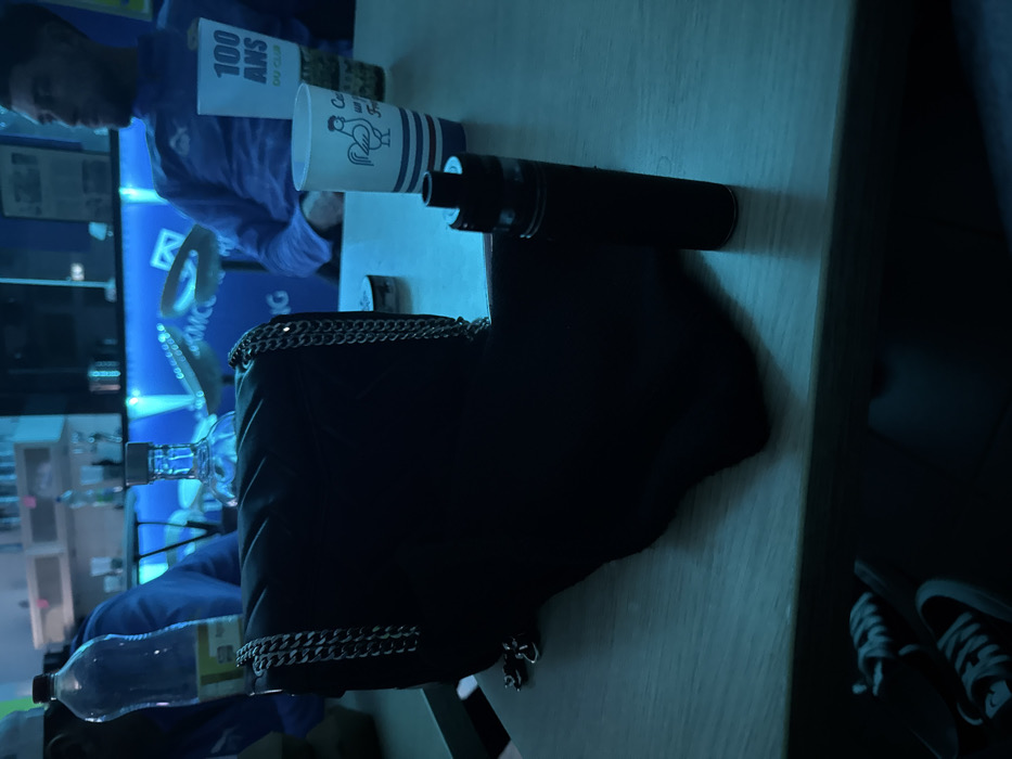 A table illuminated by blue light displays several items. A vape pen lies next to a white cup featuring a rooster design. A large black bag with silver chain detailing dominates the scene, partially obscuring a water bottle. A person in a blue shirt is visible in the background. A pair of black and white sneakers rests on the floor near the table.