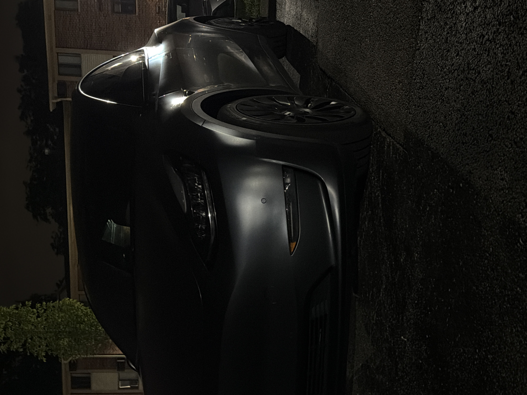 The image shows a dark-colored Tesla parked at night in what appears to be a residential area, possibly an apartment complex in Vernon Hills, United States of America. The car is the primary subject, with its front and side visible. The lighting is dim, with some artificial light sources reflecting off the car's surface, highlighting its curves and the headlights. In the background, there are parts of buildings and trees. The ground is asphalt, possibly a parking lot or a street. There are no people or any other vehicles clearly visible in the frame.
