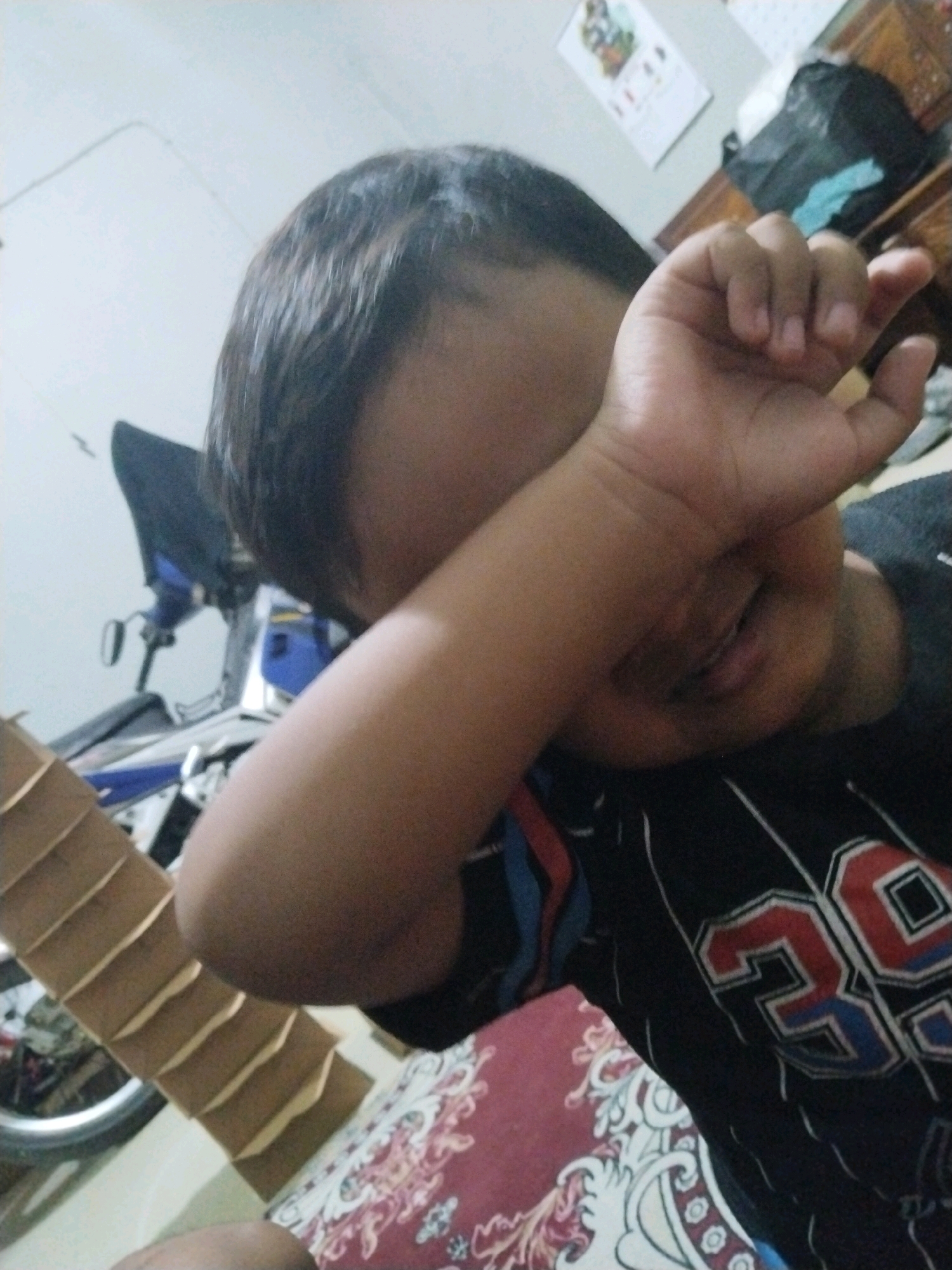 A close-up, low-angle, indoor shot shows a young boy covering his face with his right arm. He appears to be wearing a black t-shirt with red and blue accents and the number "39" printed on it. His hair is dark and short. In the background, a motorcycle with blue accents is visible. To the left, a stack of brown cardboard boxes is present. The floor is covered with a patterned rug. The overall lighting suggests a daytime setting. The image is likely taken in Pepelegi, Indonesia.