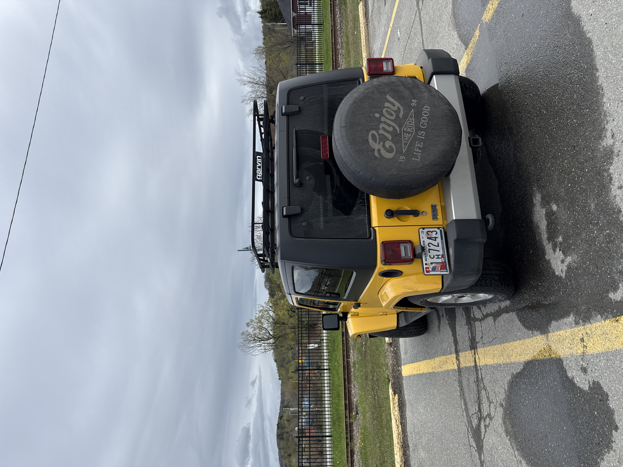 A yellow Jeep is parked diagonally in a parking space demarcated by yellow lines, with its rear facing the camera. The vehicle has a black hardtop and a tire cover that reads "Enjoy 1994 The Ride, Life is Good." A "Garvin" rack is mounted on the roof. The Maryland license plate number H7243 is visible. Behind the car is a black metal fence bordering a grassy area with trees. The sky is overcast. The location appears to be Newport, United States of America based on the metadata.