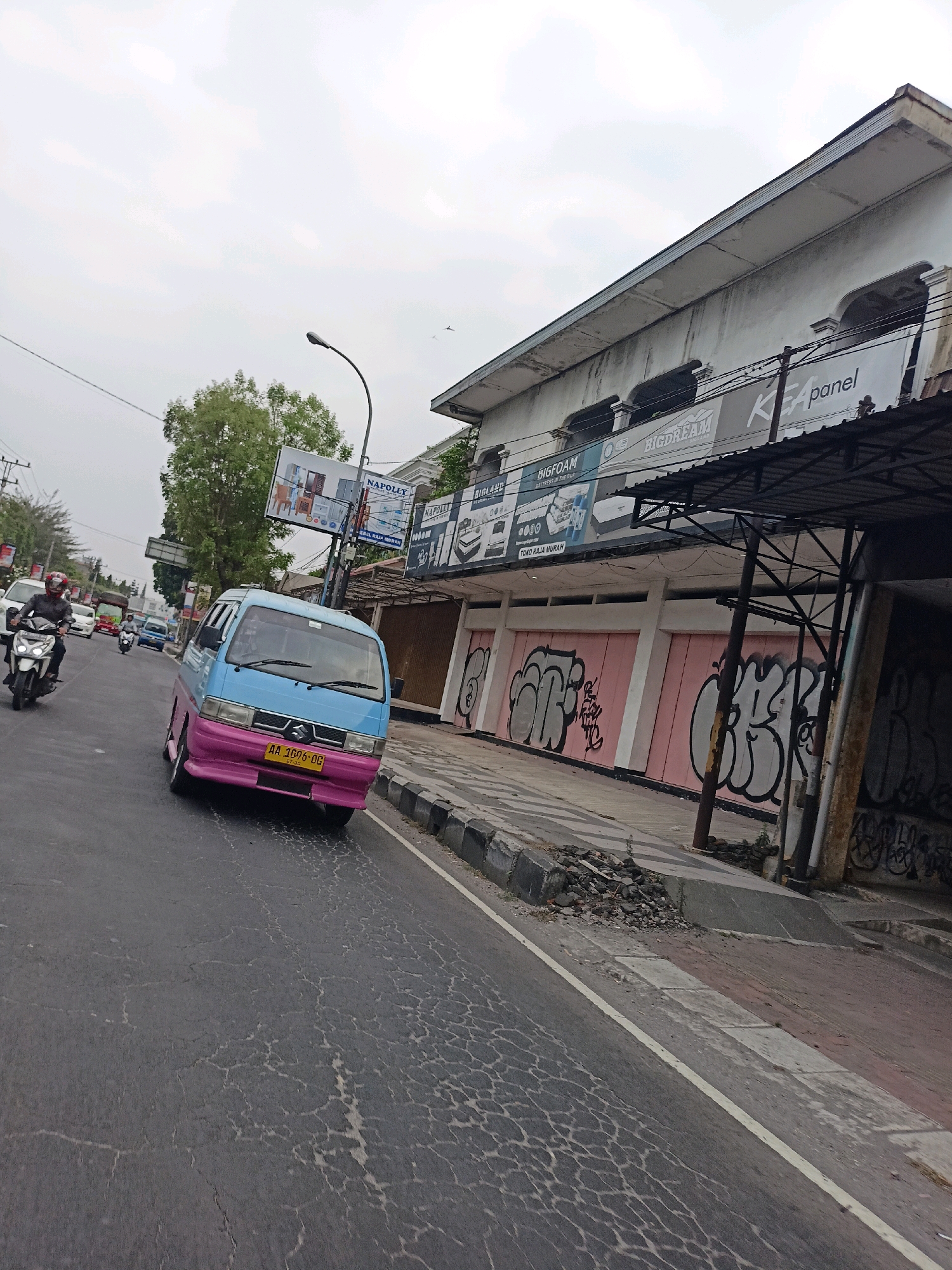The image shows a street scene in Magelang, Indonesia. In the foreground, a light blue and pink van is driving on a cracked asphalt road. A motorcyclist wearing a helmet is seen in the background on the left. To the right of the road, there is a building with advertisements on its facade, including "BIGFOAM," "BIGDREAM," and "KEA panel." The building's pink storefront is covered in graffiti. The sky is overcast.