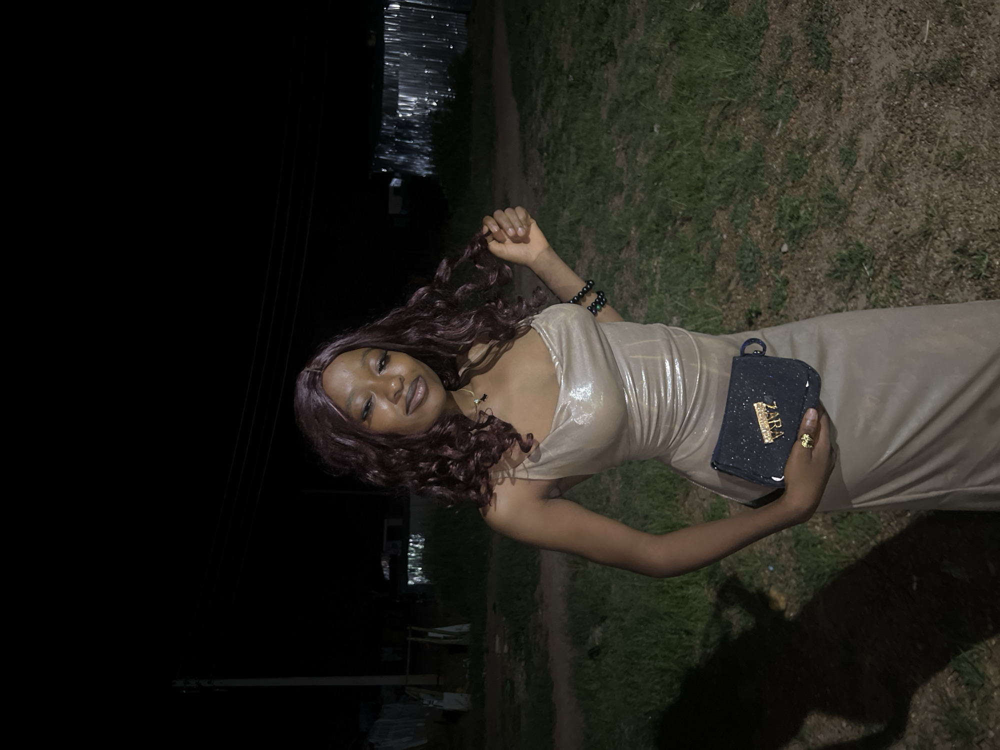 A young Black woman with dark, curly hair and dark skin is standing outdoors at night. She is wearing a shimmering, champagne-colored slip dress and a dark blue clutch purse with gold lettering. She has dark jewelry on her wrist and a gold ring on her finger. Her left hand is holding a strand of her hair. The background is dark with some distant lights and structures, possibly in the city of Durumi, Nigeria. The ground is covered in grass and dirt. A shadow of the woman is cast on the ground to her left.