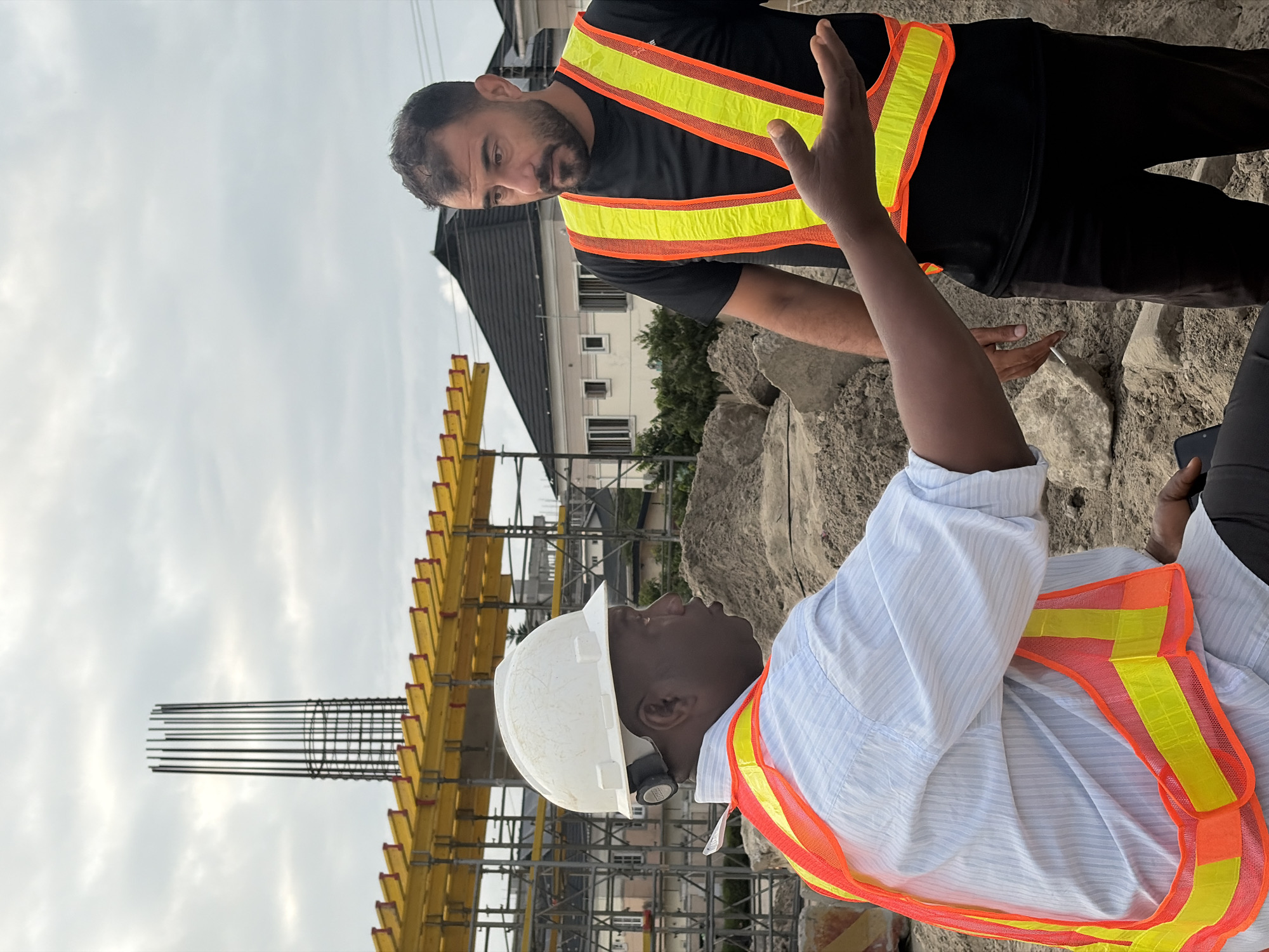 Two men in high-visibility vests are talking at a construction site in Alaguntan, Nigeria. The man on the left, wearing a white hard hat and a light blue striped shirt, is gesturing with his right hand as he speaks. The man on the right, wearing a black t-shirt, is looking at the speaker. In the background, a concrete pillar reinforcement cage stands tall against a cloudy sky, with yellow scaffolding visible below it. Residential buildings and trees are also present in the distance, indicating an urban or suburban setting. The ground is covered with dirt and rocks, typical of a construction environment.