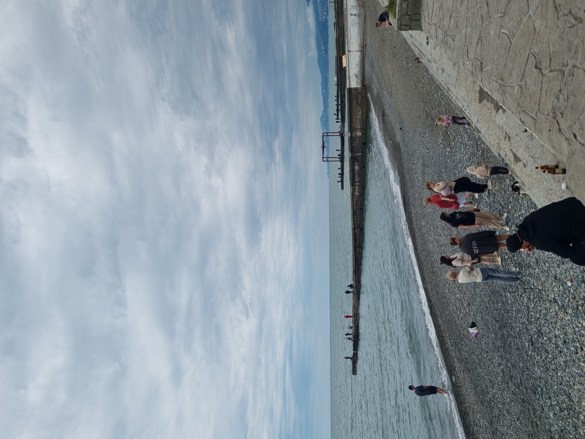 The image shows a pebble beach in Sochi, Russia. The sky is cloudy. A concrete walkway borders the beach, which is lined with people. Some people are standing and looking at the sea. The water is gray-green. Further along the beach, a pier extends into the water with a few people standing on it. In the distance, mountains are visible.