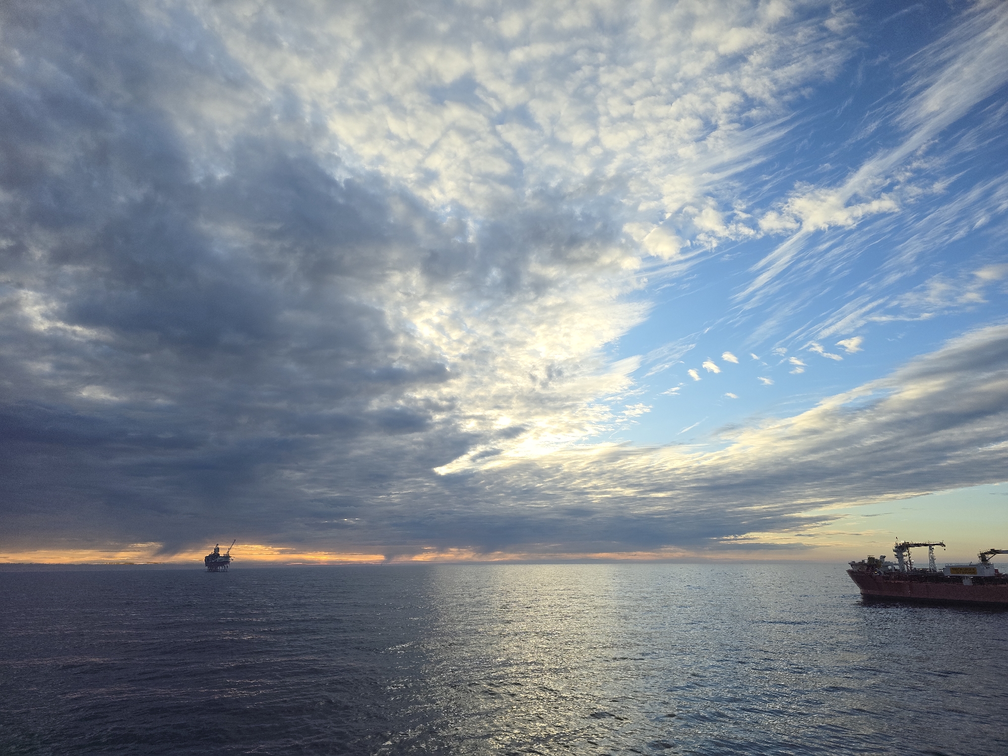 The image depicts a vast expanse of ocean under a dynamic sky. On the left horizon, a platform structure stands silhouetted against a band of orange-tinted light. A vessel is visible on the right side of the frame. The sky above is a mix of dark, heavy clouds on the left, transitioning into lighter, wispy clouds and patches of blue on the right. The ocean's surface reflects the ambient light, creating a shimmering effect. The city location is not discernible from the image.