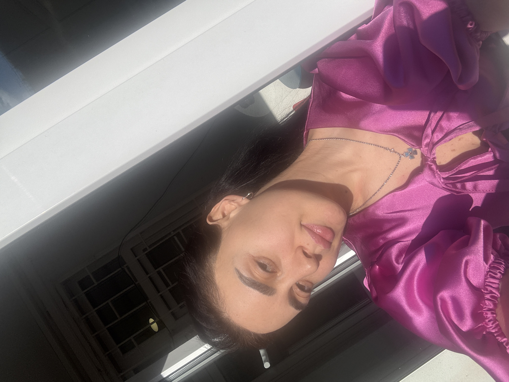 A woman with dark hair, wearing a purple satin top and a silver necklace, is standing by a window. The window has a white frame and metal security bars. The woman is smiling and looking at the camera. The image is taken in Tbilisi, Georgia, as indicated by the architecture visible through the window.