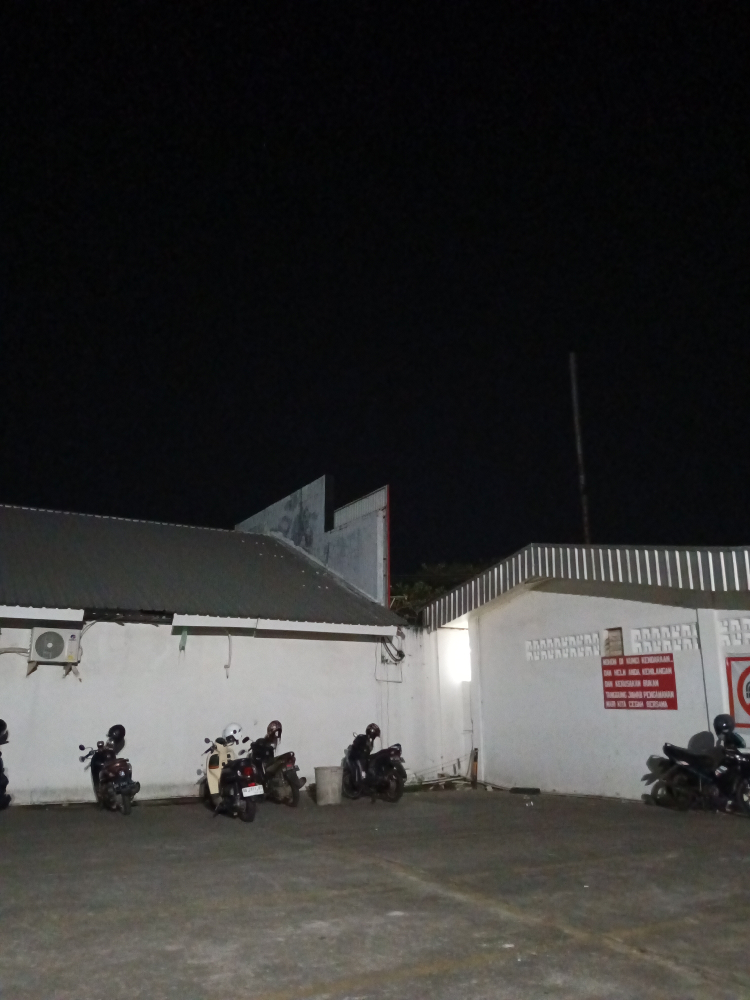 This is a nighttime photograph of a building facade and an outdoor parking area. The building is white with a dark, corrugated metal roof. An air conditioning unit is mounted on the left side of the wall. Several motorcycles are parked in the foreground, some with helmets on their seats or handlebars. A red sign with white text is visible on the wall of the building on the right side. The ground is paved with asphalt. The sky is completely dark, indicating it is night. Based on the signage and common infrastructure, this location is likely a commercial or industrial building in Makassar, Indonesia.