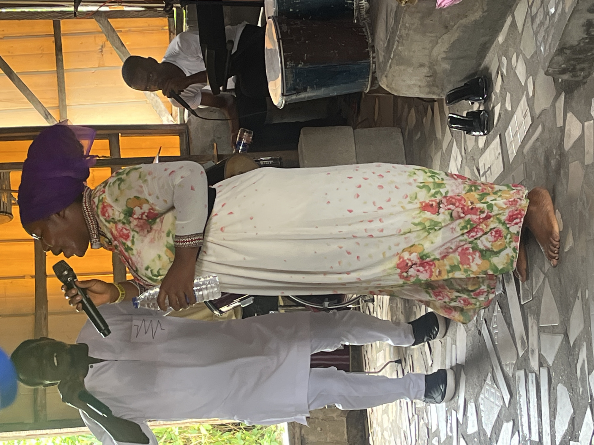 The image shows a group of people under a shelter in Iranla, Nigeria. An African woman with a purple head covering, wearing a floral dress and multiple necklaces, is speaking into a microphone while holding a water bottle. Below her, a man in a white outfit is also speaking into a microphone. Behind them and slightly above, a man in a white shirt is speaking into a microphone while sitting near a drum. A pair of black shoes is on the patterned floor near the woman. The floor and wall behind the drum are concrete. The shelter has a wooden roof.