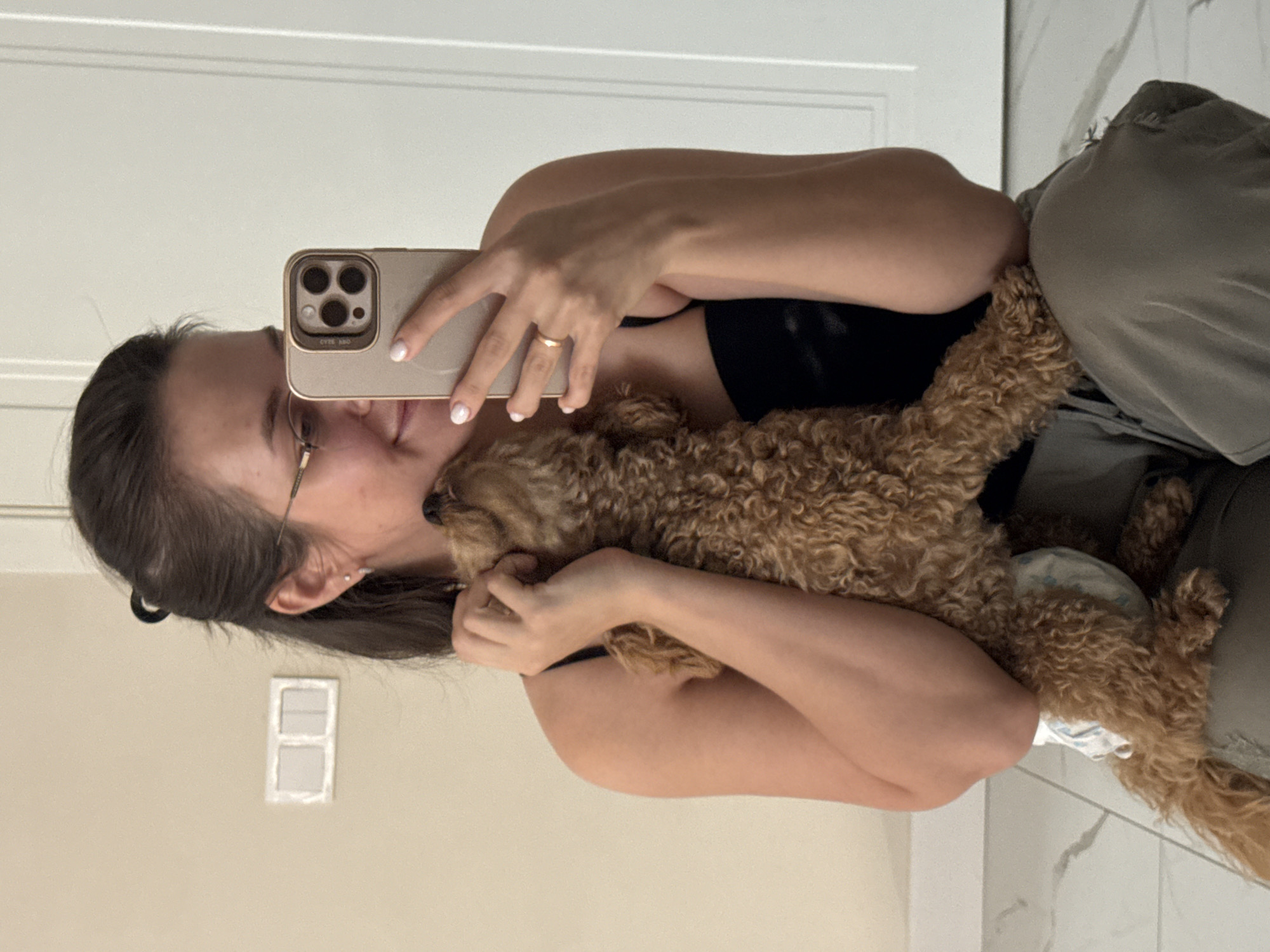 A young woman with dark hair pulled back and glasses takes a selfie while holding a small, light brown poodle. The poodle is wearing a diaper and is snuggled against the woman's chest. The woman is wearing a black tank top and grey pants. She has a ring on her left hand. The background appears to be an indoor residential setting, possibly a hallway or room with light-colored walls and a white door. The floor is tiled with a marbled pattern. There is a light switch on the wall to the left of the woman. This scene is not in Pervomaysky, Russia, as it is an indoor domestic setting without any identifiable Russian landmarks or context.