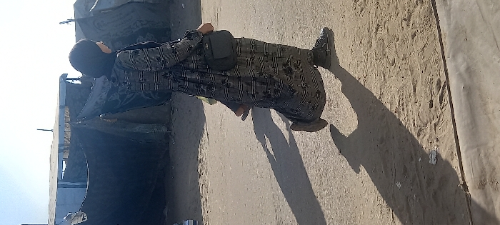 A solitary individual, appearing female, is captured mid-stride walking from right to left on a path in Khan Yunis, Palestinian Territory. The individual wears a dark hijab, a long dark-patterned abaya or dress, and a dark shoulder bag positioned across the body. The left foot is lifted, revealing a dark shoe, while the right foot is on the sandy ground. A prominent, elongated shadow is cast forward on the dusty and partly paved surface. In the background, a light blue, low-lying structure is visible. Adjacent to and partially behind this structure are several dark, makeshift shelters constructed from tarpaulins or fabric, indicative of a temporary settlement. The sky overhead is clear and bright.
