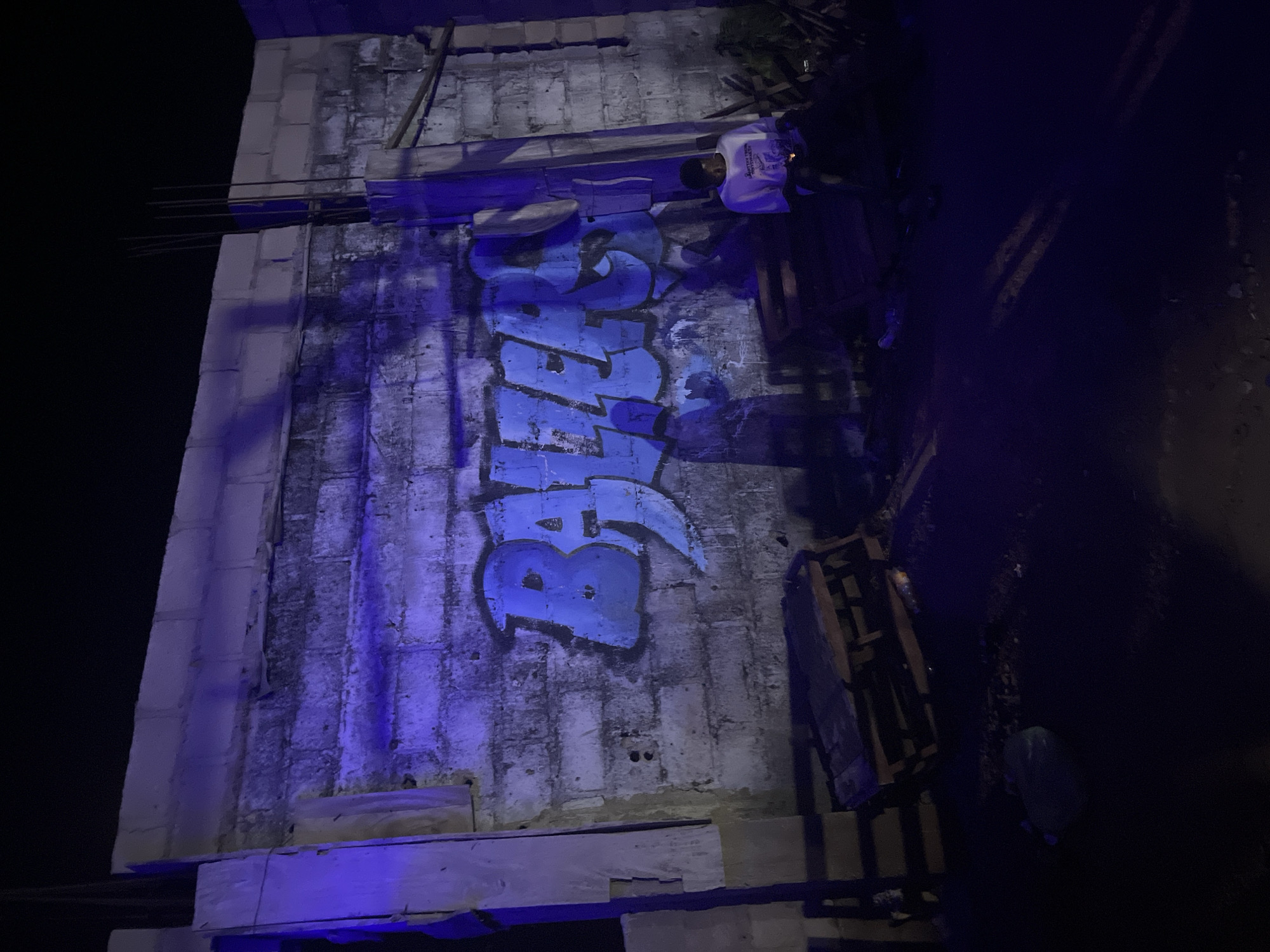 The image captures a night scene in Durumi, Nigeria, illuminated by a harsh blue light, likely from an artificial source. A rough-textured concrete block wall dominates the frame, bearing large, blue graffiti that appears to spell out "BALERS". The wall shows signs of wear and tear, with visible mortar lines and uneven surfaces. To the left of the graffiti, a section of lighter colored, unfinished wall is visible. In the foreground, several wooden pallets are stacked haphazardly. To the right of the graffiti, a young person, appearing to be male, is seated on a dark surface, wearing a white t-shirt with a graphic print and dark pants. This individual is looking downwards, holding a small object in their hands, and is bathed in the blue light. Shadows are cast across the scene, adding to the overall low-light ambiance. The ground appears to be unpaved, with scattered debris. The background is predominantly dark, suggesting it is nighttime. The overall impression is one of an outdoor urban or semi-urban environment, possibly an industrial area or a construction site, given the materials and unfinished construction elements.