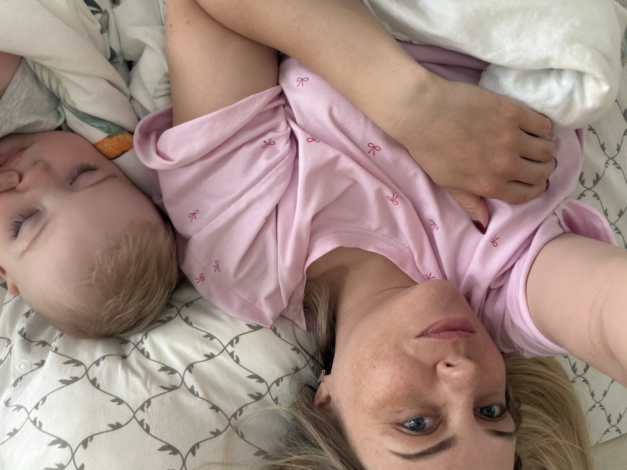 A woman and a baby are lying on a bed. The woman is wearing a pink shirt with small bow designs. The baby is lying next to her, eyes closed, with light blonde hair. They are both lying on a white patterned sheet, and there is a white blanket bunched near the woman's arm. The scene appears to be indoors, possibly in a home. There are no explicit clues to suggest a specific location in Votkinsk, Russia. The image mainly depicts a mother and child resting.