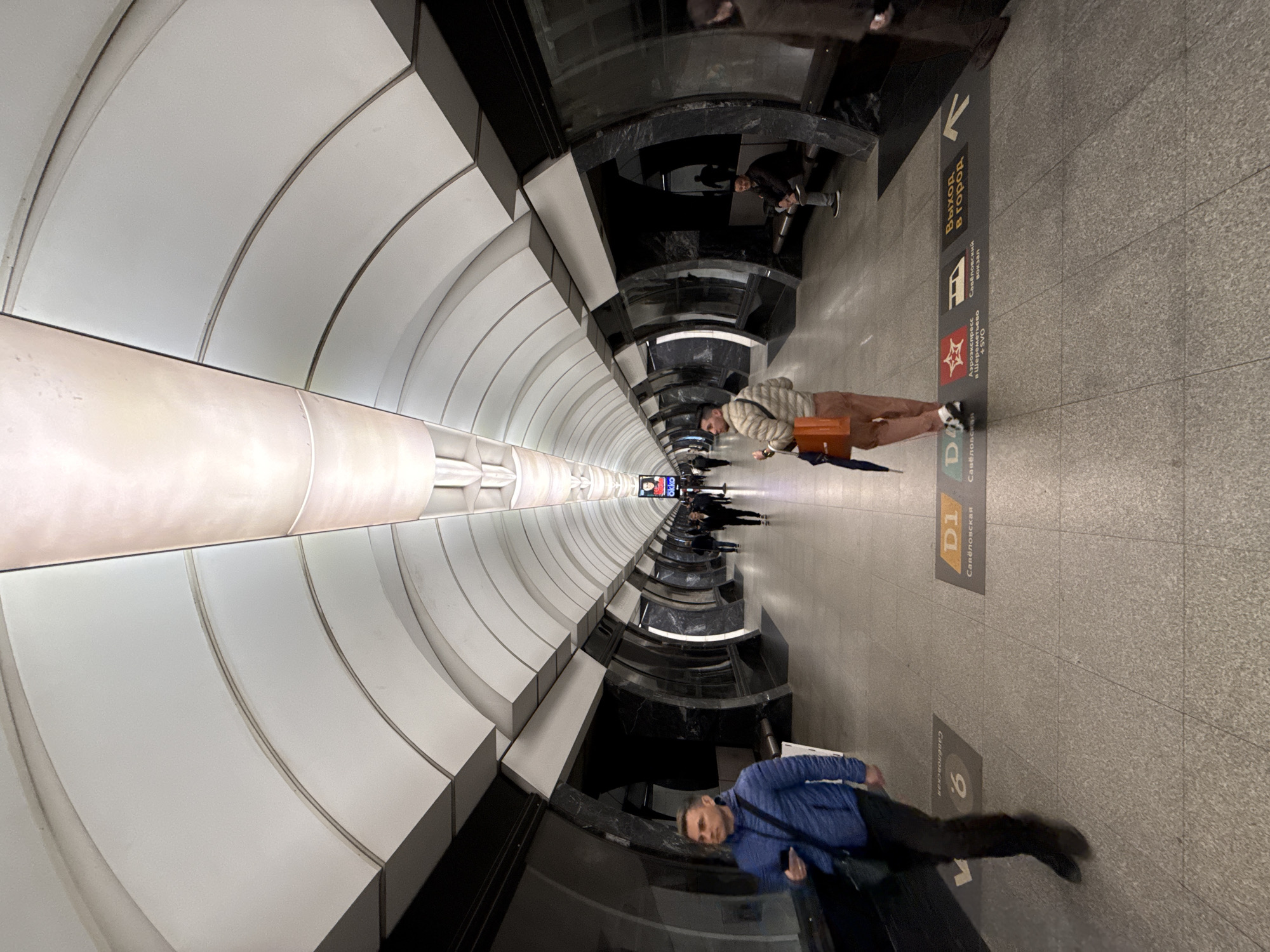 The image shows the interior of a Moscow Metro station, specifically the Savyolovskaya station, as indicated by the signage on the platform floor. The station has a unique architectural design with a long, arched tunnel illuminated by a central lighting fixture. Two individuals are visible: one wearing a blue jacket, walking towards the camera; and another with a tan jacket, standing still carrying a satchel. The flooring is gray tile, with signs indicating directions, including "Exit to the city," and the route to Sheremetyevo Airport. Other people are visible in the background, further down the platform. The environment is clean and well-lit.
