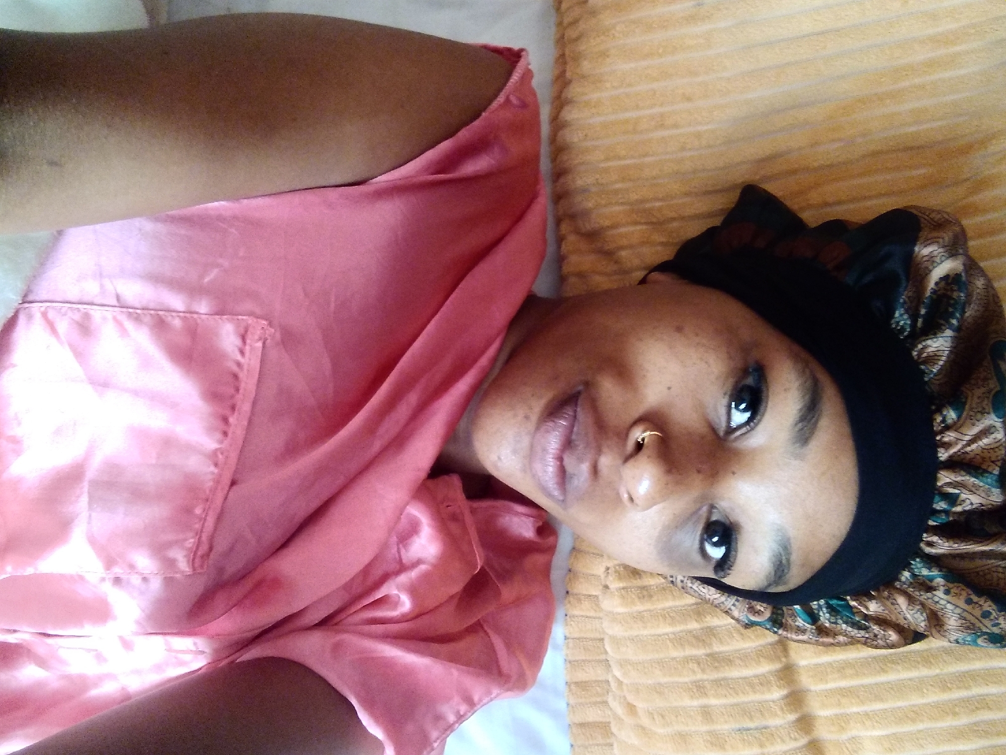 A close-up, overhead shot shows a young Black woman lying down, wearing a satin bonnet with a paisley pattern and a pink satin pajama top. She is resting on a yellow, striped pillow. Her face is turned towards the camera, with her eyes open and her mouth slightly parted. A small gold hoop pierces her left nostril. The background consists of the pillow and a white surface, likely a bed. The scene is indoors, possibly in a residential setting in Nyeri, Kenya.
