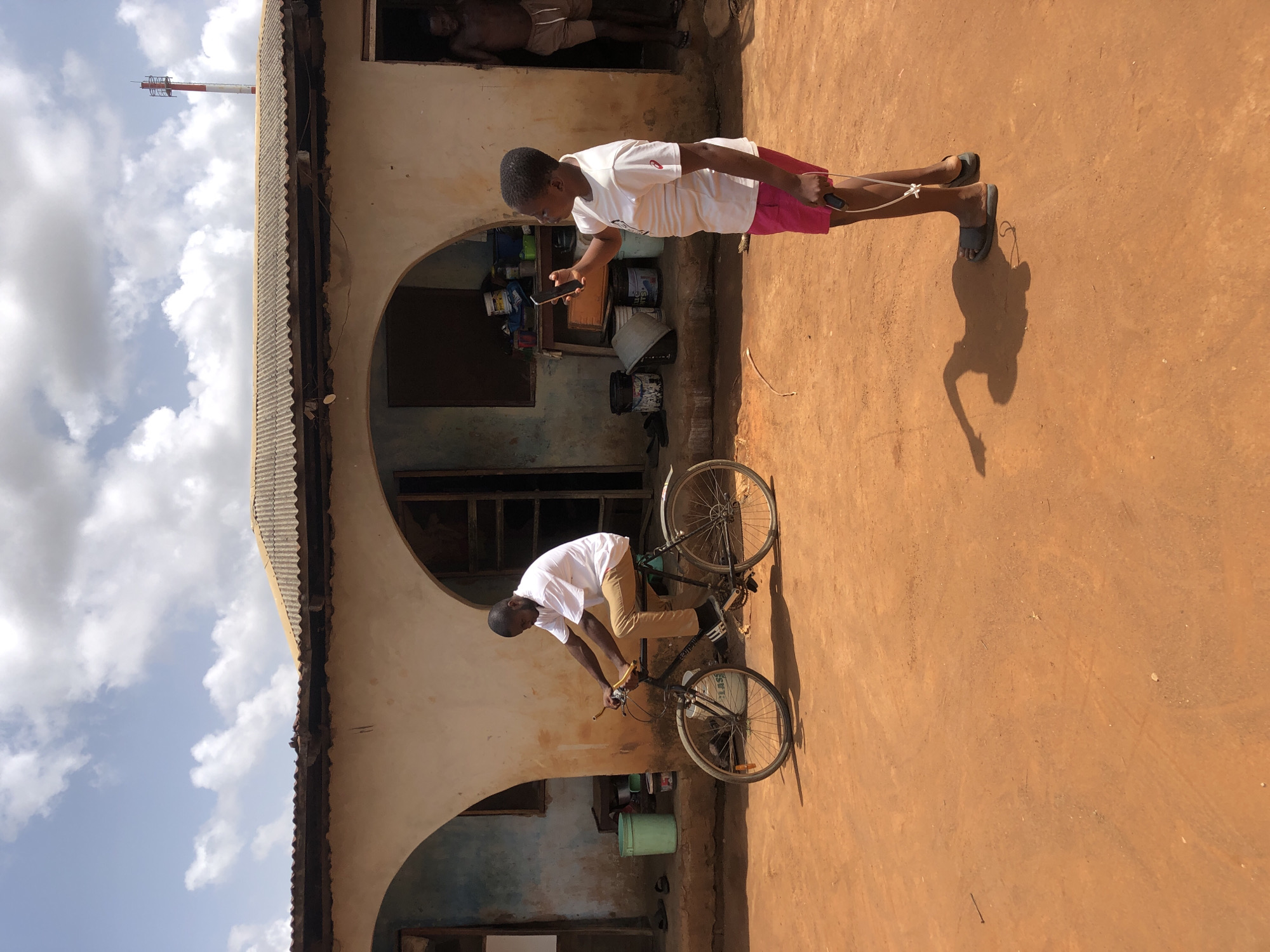 Two people are captured near a building with arched entryways. One is on a bicycle and another is using a cell phone. A partial image of a communication tower is in the background. The location is in the city of Unknown, Unknown.
