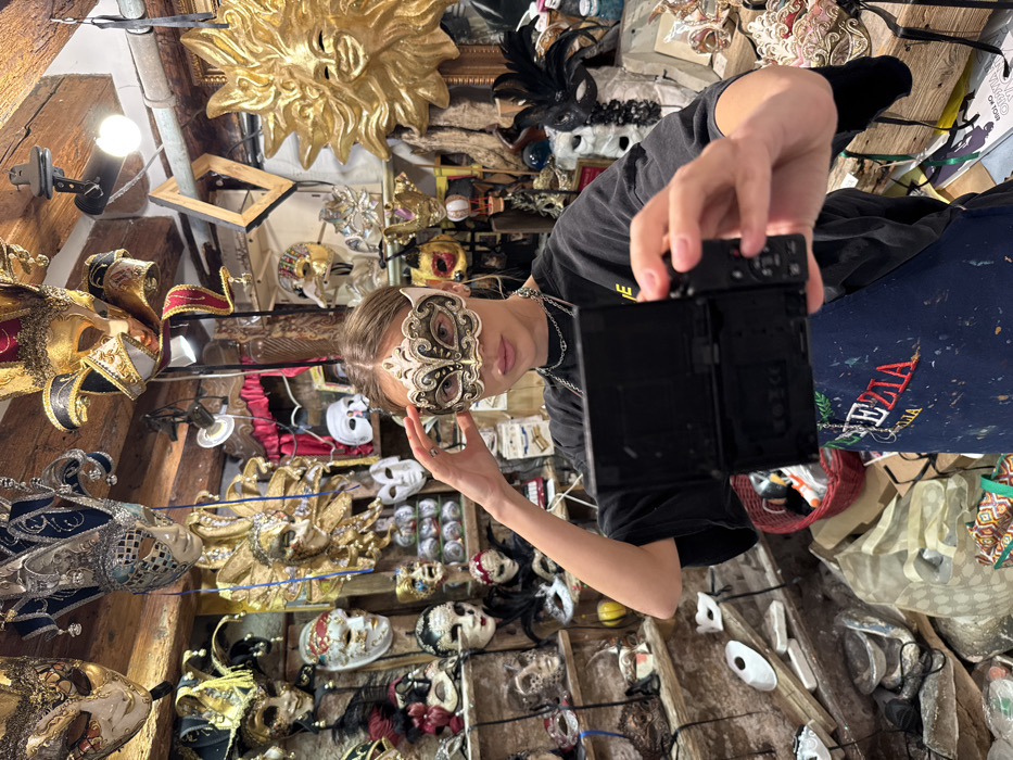 A young adult female is depicted in a traditional Venetian mask shop or artisan workshop located in Venice, Italy. She is wearing an ornate, predominantly white and gold half-mask over her eyes and nose, featuring intricate dark scrollwork. She holds a black digital camera in her right hand, pointed towards herself, indicating a self-portrait capture. Her left hand is positioned near the left side of the mask. She wears a black short-sleeved top and a dark blue apron with "VENEZIA" embroidered in red, along with multiple silver chain necklaces. The shop's interior is densely packed with a wide variety of Venetian masks of different styles, colors, and sizes, displayed on walls, suspended from the wooden-beamed ceiling, and arranged on shelves. A large, golden sun-faced mask is prominently featured on the right side of the frame. The lower sections of the shop display additional masks and general clutter consistent with an active artisan workspace or retail environment.