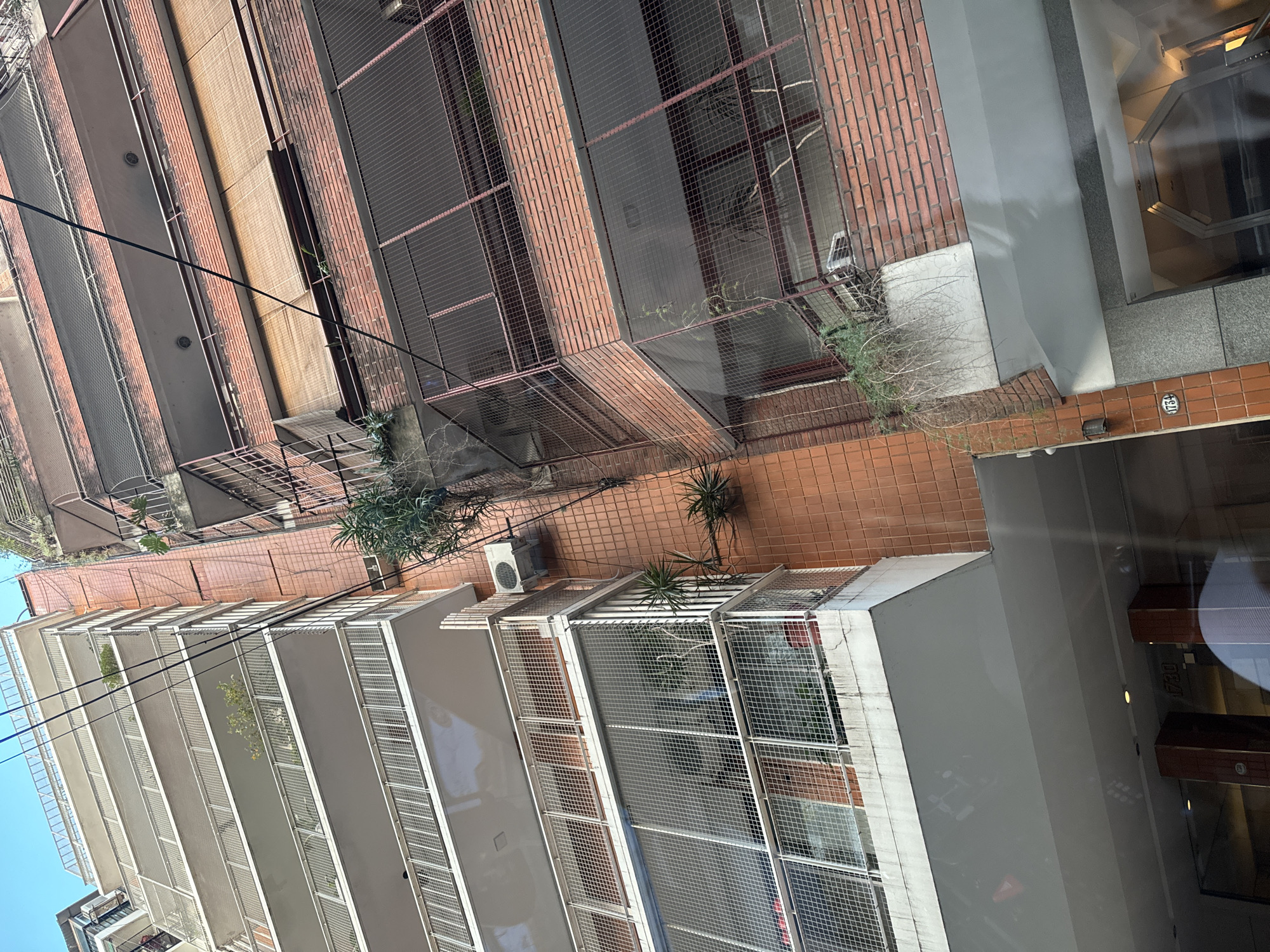 The image captures an outdoor urban scene, likely from an elevated position, showcasing architectural details of adjacent buildings.
Building features:
A red brick building with a metallic grid façade is visible on one side. An adjacent white-colored building with similar grid balconies is present. There is an air conditioning unit.
Address:
There is an address, "1734", marked on a wall.
Vegetation:
Sparse greenery includes plants on balconies and near the building foundations.
Location:
Given the street number format and architectural style, the image is potentially situated within Buenos Aires, Argentina.