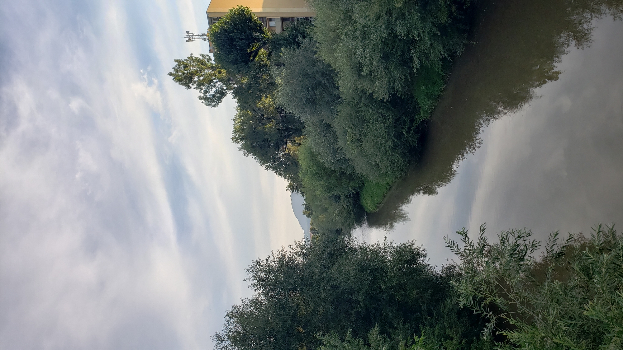 The image captures a tranquil scene of the Torysa River flowing through a vegetated urban environment in Prešov, Slovakia. Lush green trees and bushes line both banks of the river, with the foliage creating a natural corridor. The water reflects the cloudy sky and the surrounding greenery. In the background, a gentle hill is visible, adding to the scenic backdrop. On the right bank, a light-colored building with windows and a telecommunications tower on its roof stands amidst the trees, indicating the presence of urban infrastructure. The sky is partially cloudy with streaks of white clouds against a pale blue backdrop. The overall atmosphere is peaceful and serene, showcasing a harmonious blend of nature and urban elements.