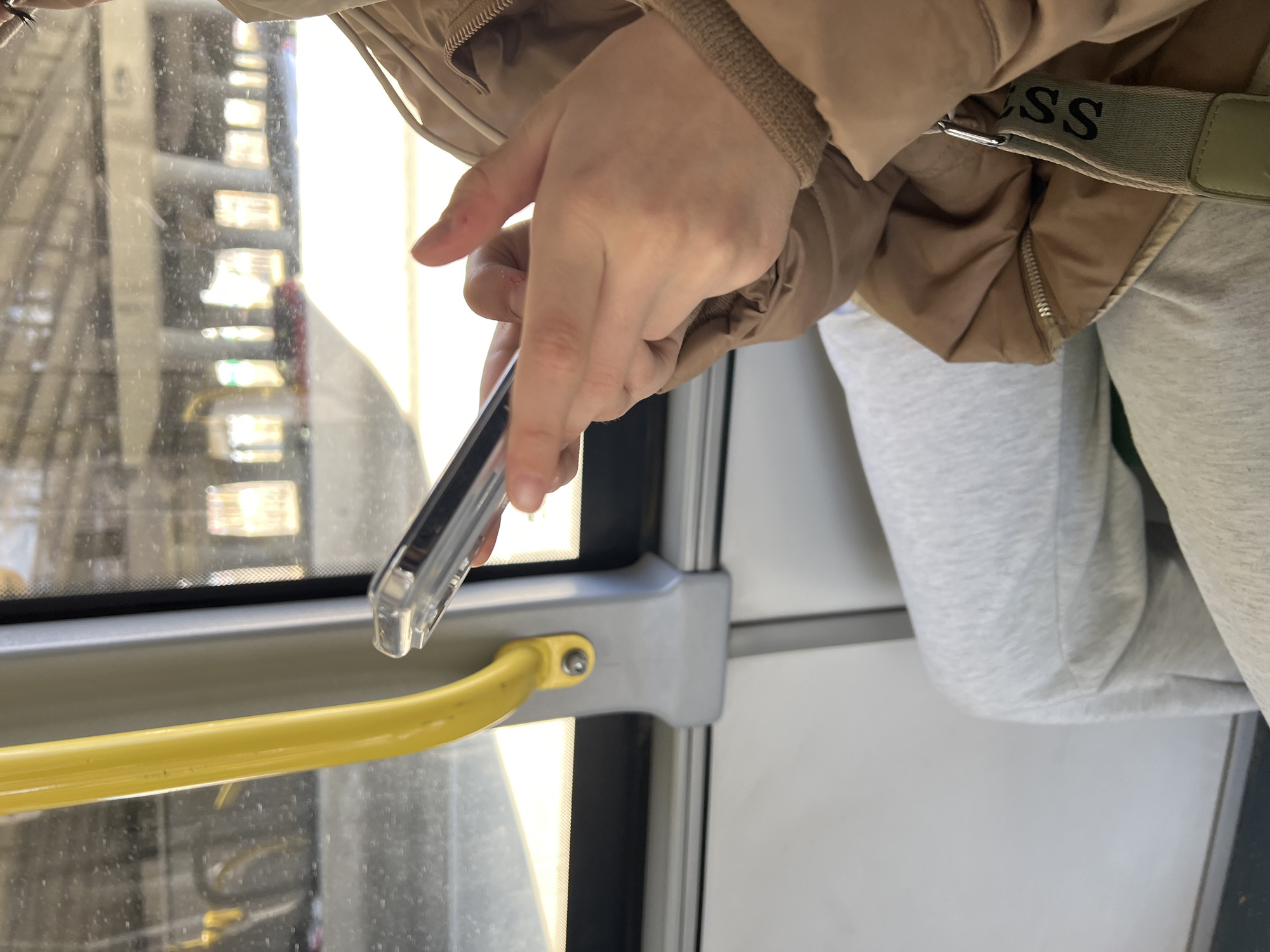 A person wearing a beige jacket with a "Guess" logo on the strap and grey sweatpants is holding a smartphone in their hand. The person is inside a bus or tram, next to a window with a yellow handrail and a grey frame. The view outside the window appears to show a building with many windows. The location is likely Tyumen, Russia.