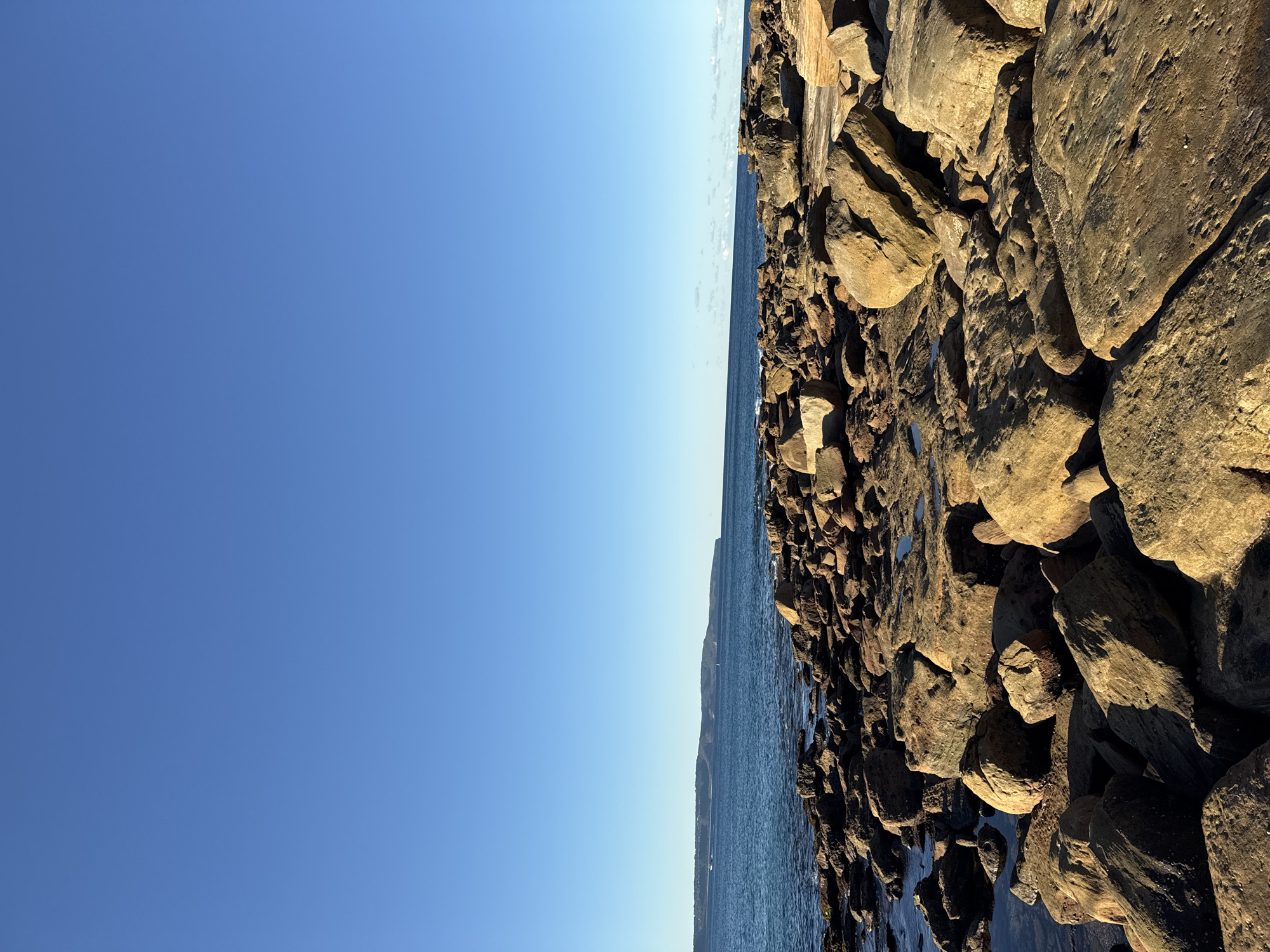 The image depicts a rocky shoreline on a clear, sunny day. In the foreground, numerous large, weathered rocks dominate the scene, scattered across the land and reaching towards the water. Some of these rocks have a golden-brown hue, illuminated by the sunlight. The water of the ocean is visible in the midground, a deep blue color, with small waves lapping against the shore. In the background, a distant landmass with rolling hills can be seen, also under the clear blue sky. There are no people or any man-made structures visible in this particular frame. The environment appears natural and untouched, showcasing a rugged coastal landscape. The location is identified as the city of Palm Beach, Australia.