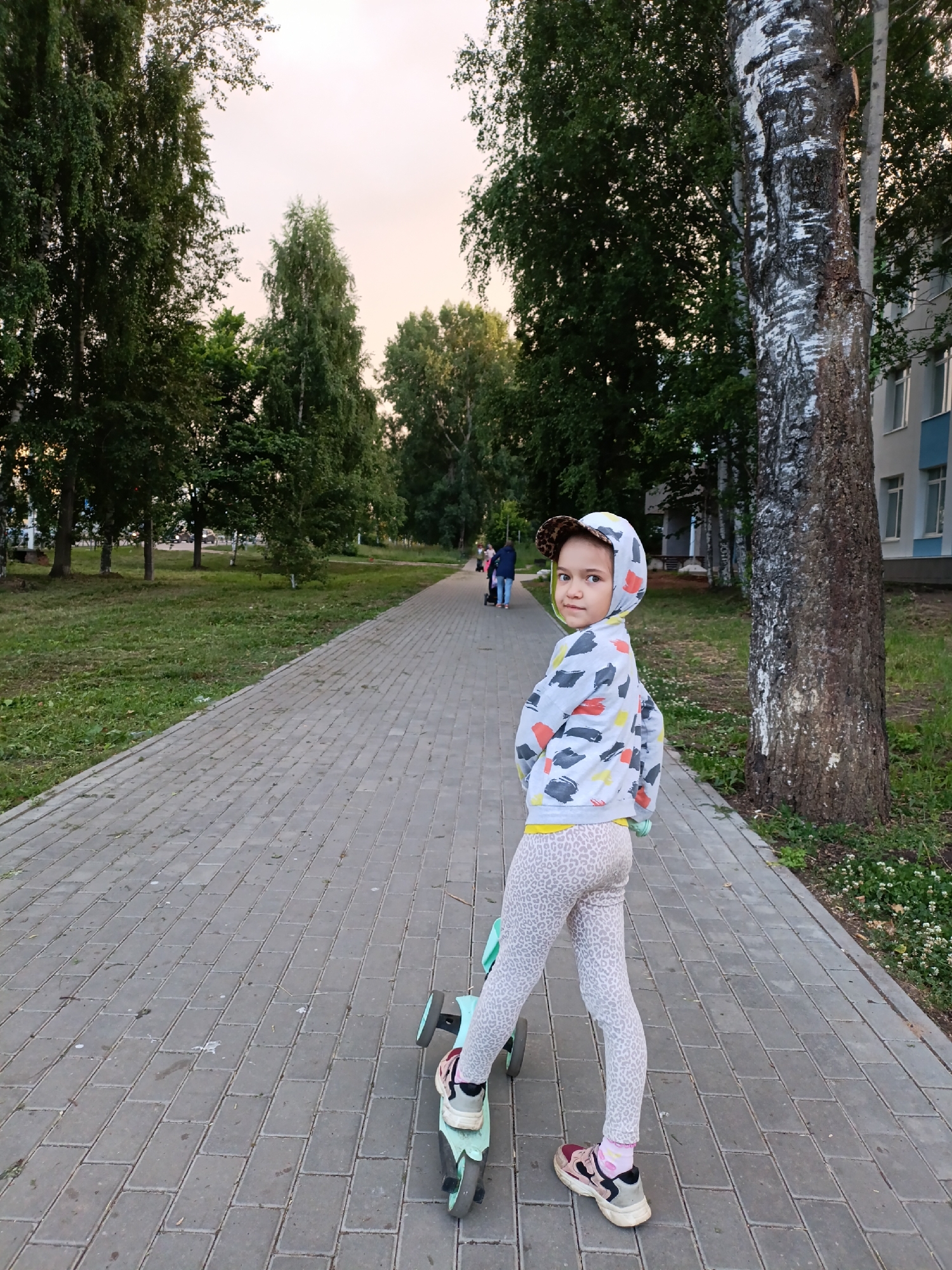 A young girl, approximately 4-6 years old, stands on a paved pathway with a light blue scooter. She is looking over her left shoulder at the camera with a slight smile. She is wearing a white hooded sweatshirt with a colorful abstract print, leopard-print leggings, pink and white socks, and athletic shoes. A baseball cap with a leopard print pattern is on her head.
The pathway is lined with green grass and mature trees on both sides. In the background, other people are walking and some are pushing strollers. A multi-story building with light blue and white siding is visible on the right side of the frame, behind a large birch tree. The sky is overcast with a soft, peachy glow, suggesting it is either early morning or late evening. The scene appears to be a park or a residential area with a designated walking path. The overall mood is relaxed and casual, typical of a pleasant outdoor activity.