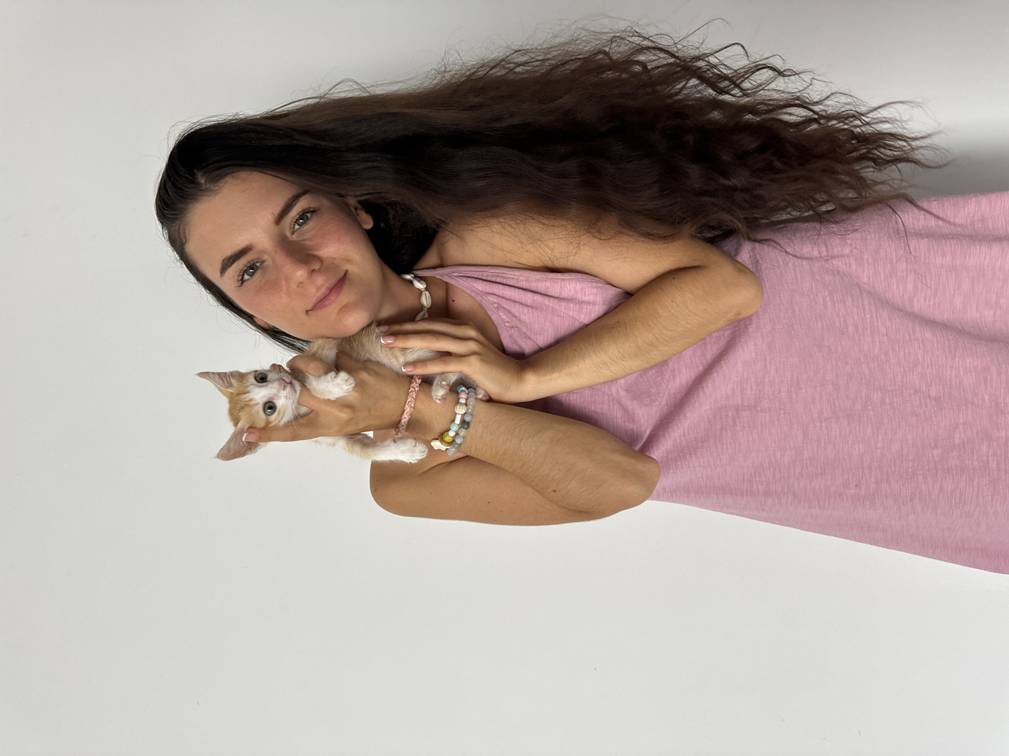 An intimate portrait captures a young woman gently holding a small kitten against a plain white background. The image is rotated 90 degrees clockwise, presenting the woman lying on her side, looking directly at the viewer with a warm, soft smile. She has long, flowing dark brown hair with natural waves, light skin, and dark eyes.
The woman is wearing a simple, sleeveless pink top or dress. Around her neck, she sports a necklace made of numerous small, light-colored shells. Her right wrist is adorned with multiple bracelets: one appears to be braided in pink, while others are beaded with various pastel colors. Her left arm cradles the tiny kitten, which features distinct orange and white fur markings and wide, light blue eyes, also gazing towards the camera. The kitten's small paws are visible as it is held close to the woman's chest.
The setting is starkly minimal, with a uniform white background that suggests an indoor studio or a very clean, uncluttered space. The mood conveyed is one of affection and tenderness between the woman and the kitten. There is no visible text in the image. The scene is located in Mũi Né, Vietnam.