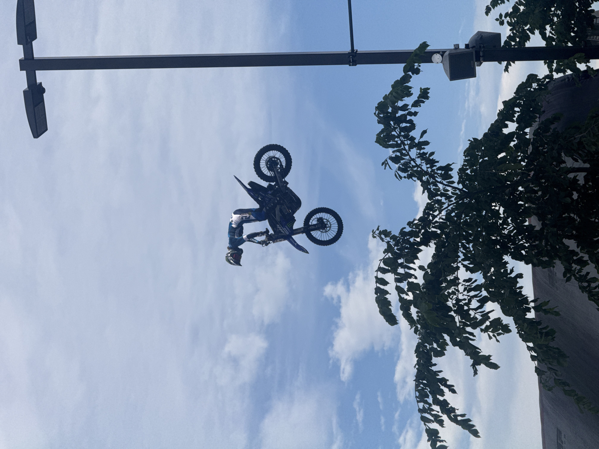 A motocross rider performs a stunt in mid-air against a cloudy blue sky. The rider is wearing a blue and white suit and a helmet, and is balanced on a blue and black dirt bike. The bike is tilted, with the front wheel pointing up. The scene is set outdoors, with the lower part of the image showing the dark green leaves of a tree. To the right of the image, a tall, dark pole with two speakers and a light fixture is visible. The background is a bright, somewhat hazy blue sky with scattered white clouds. The image appears to be taken during the daytime. There is no visible indication of the location being Calgary, Canada.