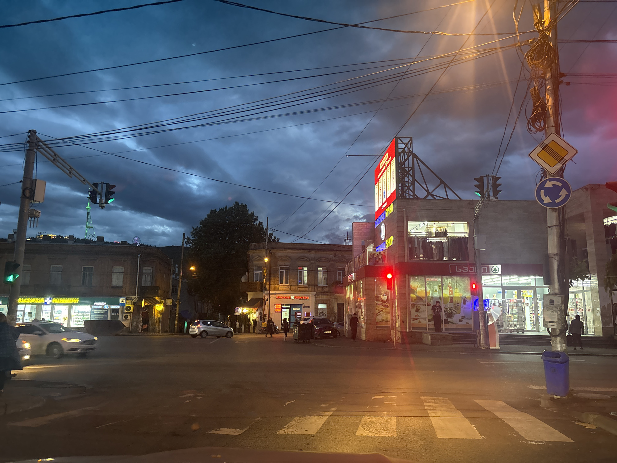 The image depicts a street intersection in Tbilisi, Georgia, at dusk. Overhead power lines crisscross above the scene. Streetlights illuminate the scene.
Buildings line the streets. A pharmacy and a bank are visible on the left side of the image. A large building with a brightly lit sign is on the right side of the frame. Traffic lights are in place at the intersection, showing red for the cross street and green for the road going away from the frame. A pedestrian crossing is visible.
Several pedestrians are on the streets. There are a couple of parked cars and a moving car in the frame. In the distance, a tower and a Ferris wheel can be seen.