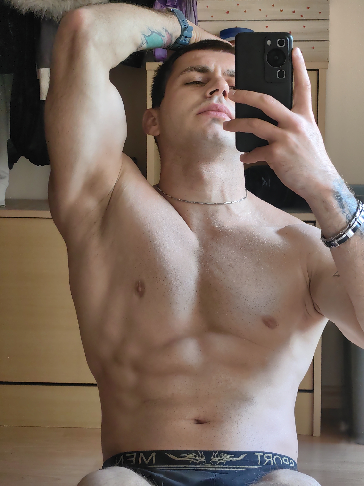 A person is taking a mirror selfie in a room. They are shirtless and wearing black underwear. The person has tattoos on their arms. The room has wooden furniture and the floor is wooden. The image is taken from the chest up.