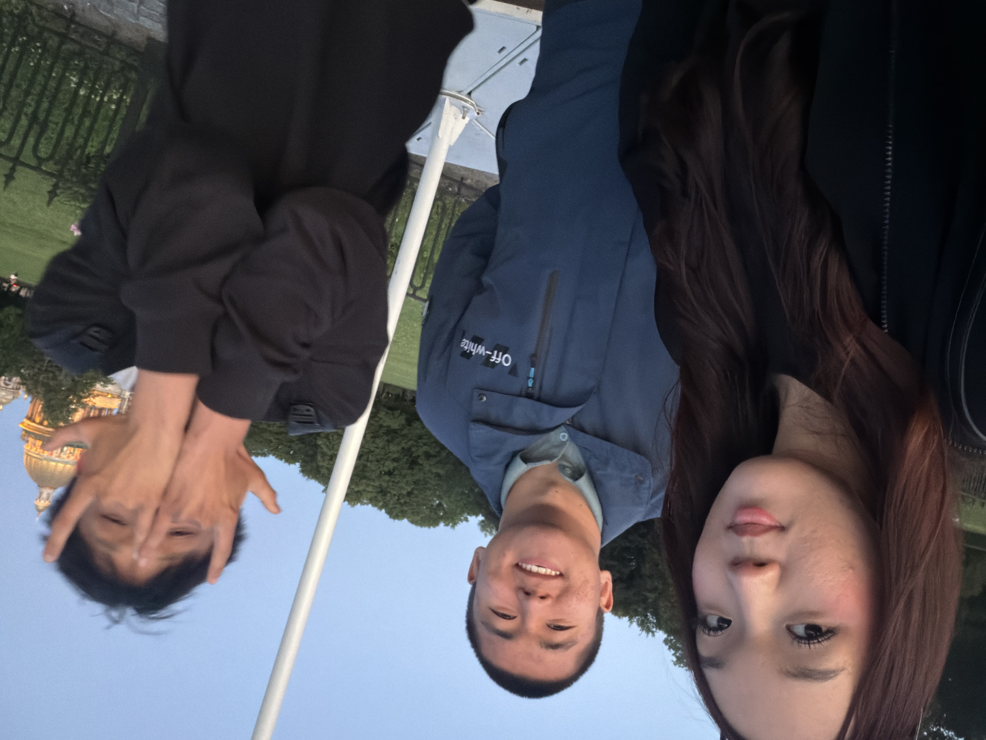 The image shows three young adults posing for a photo outdoors. The photo is taken from a low angle, with the subjects appearing upside down.
On the left is a person wearing a black jacket, with their hands covering their face. Their hair is short and dark.
In the center is a man wearing a blue jacket over a light blue shirt. He is smiling, showing his teeth. His hair is short and dark. The jacket has "OFF-WHITE" printed in white text.
On the right is a woman with long, dark brown hair. She is looking towards the camera with her eyes open and lips slightly parted. She is wearing makeup, including mascara and blush.
The background appears to be a park or garden setting with green trees and a body of water, possibly a river or canal. In the distance, a building with a golden dome and arched windows is visible, which resembles a church or government building. This dome is a landmark of Saint Petersburg. A white pole, possibly part of a flagpole or streetlamp, is also visible in the foreground. The sky is a clear blue, suggesting it is daytime, likely late afternoon or early evening given the soft lighting. The overall mood of the photo seems to be casual and happy, as indicated by the smiles and relaxed poses.