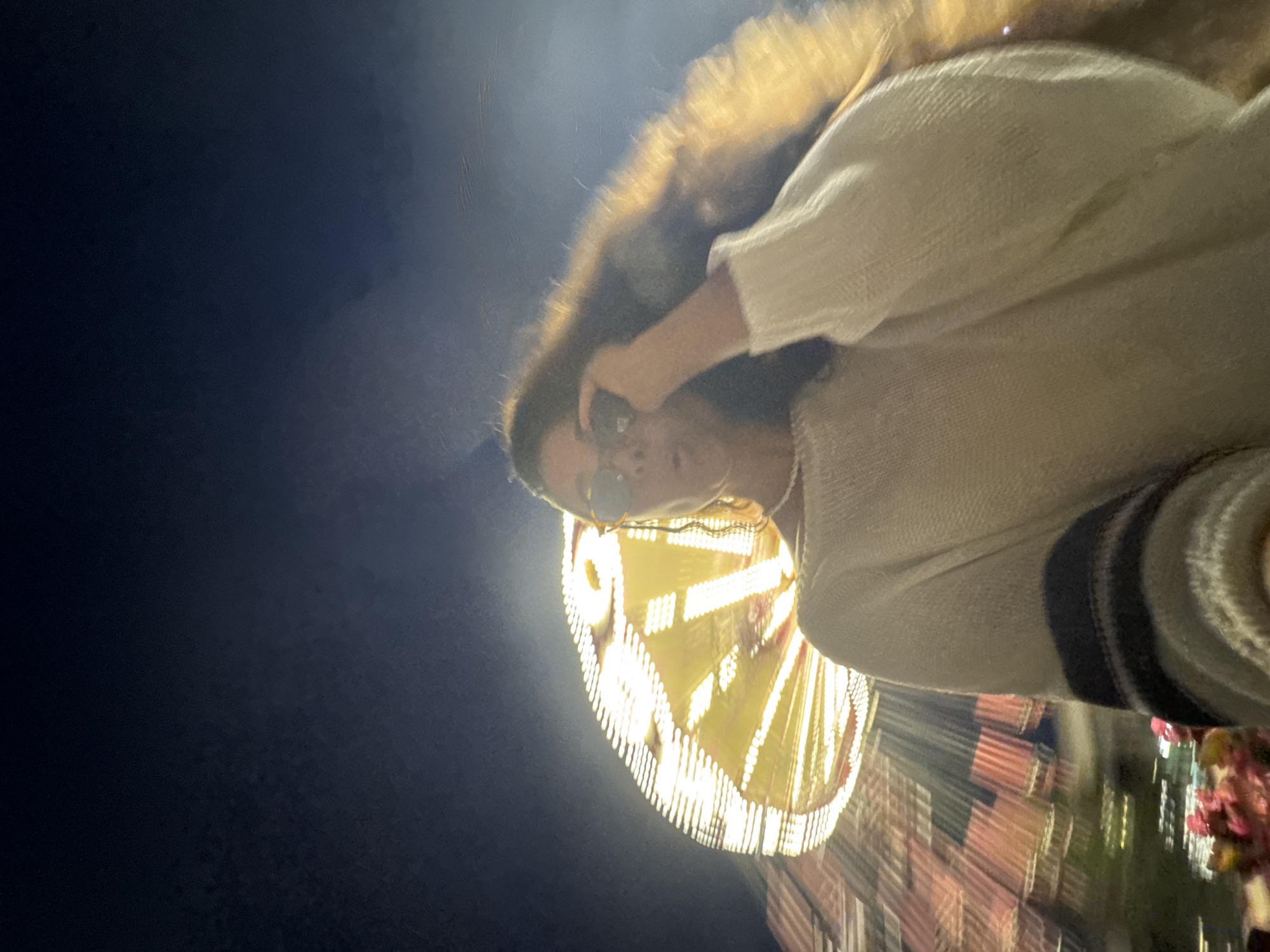 A young woman with long, wavy hair and sunglasses is taking a selfie at night. She is wearing a cream-colored sweater with striped cuffs and a pearl necklace. Behind her, a large, brightly lit amusement park ride, possibly a Ferris wheel, is spinning, causing a blur of lights and motion. The sky is dark blue, and there are some faint lights in the background. The overall impression is of a fun and exciting night out. The image is taken at an amusement park in Kazan, Russia.