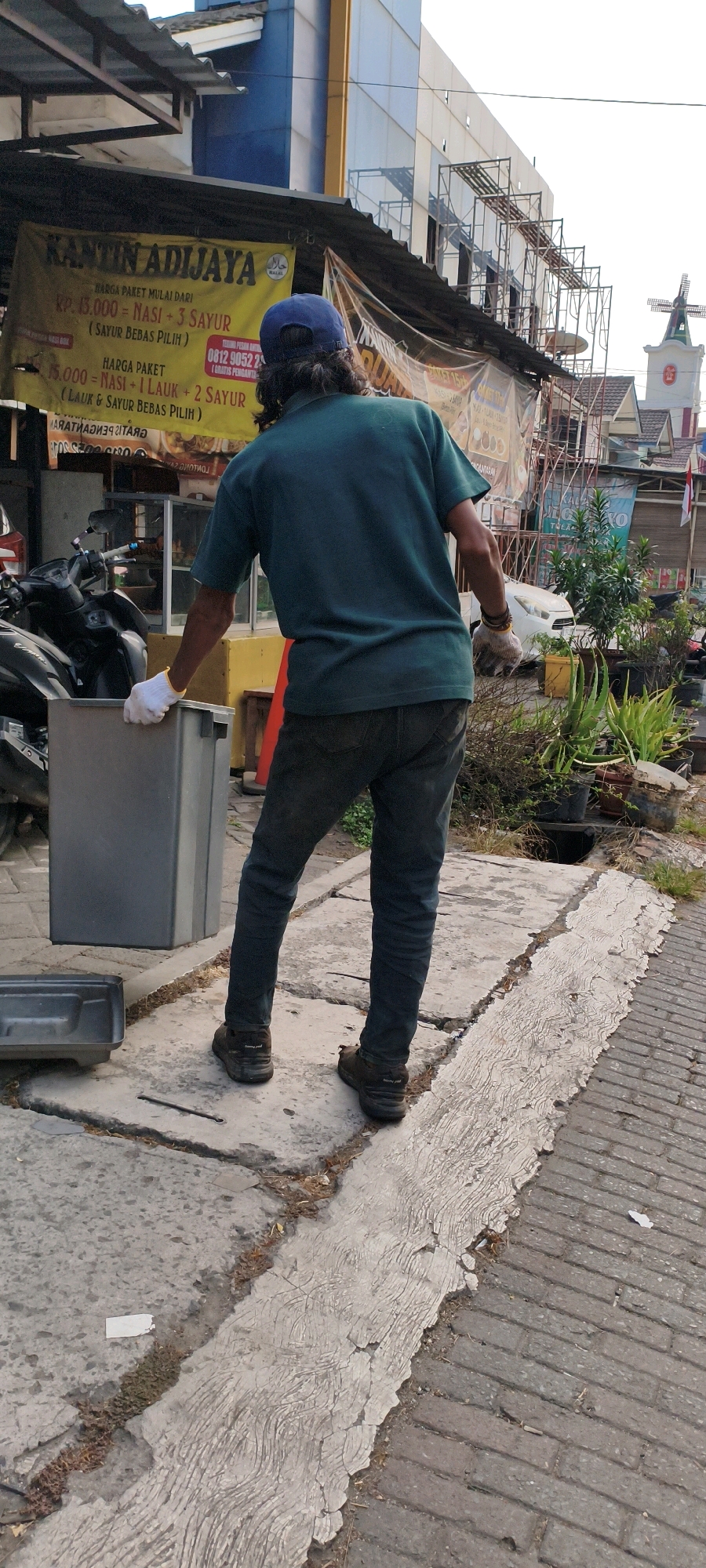 A person wearing a blue cap, dark green t-shirt, and dark denim jeans is seen from behind in what appears to be a street scene in West Jakarta, Indonesia. The person is wearing white gloves and is holding a gray plastic container. In the background, there is a banner that reads "KANTIN ADIJAYA" and lists food prices. There is also a motorcycle, scaffolding, and several potted plants. The ground is paved with concrete and bricks. The overall scene suggests a commercial area, possibly a food stall or restaurant, with ongoing construction or renovation.