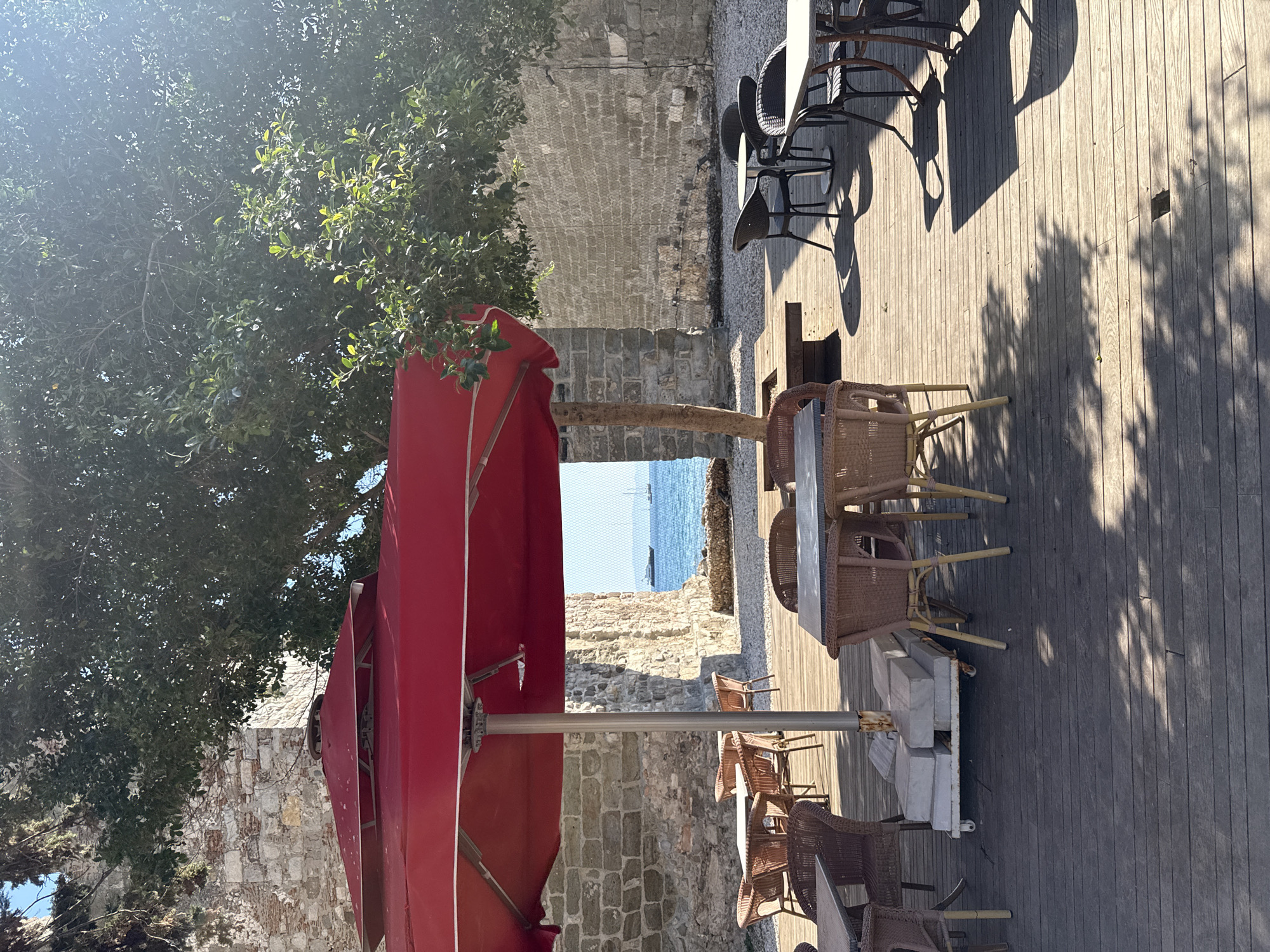 This image depicts an outdoor seating area of a cafe or restaurant located in Bodrum, Turkey, likely near the waterfront given the view of the sea and sailboats in the distance. The foreground is dominated by a wooden deck furnished with several tables and chairs. Some tables are made of woven wicker with square tops, while others have round, white tops and dark, metal chairs. A large red umbrella provides shade over some of the tables. Behind the seating area, a stone wall, possibly part of a historical structure, forms a backdrop. A large tree with dense foliage casts dappled shadows across the deck, adding to the serene atmosphere. The sea is visible beyond an opening in the stone wall, with clear blue skies above. There are no people visible in the image. The scene suggests a relaxed, sunny day at a seaside establishment in Bodrum.
