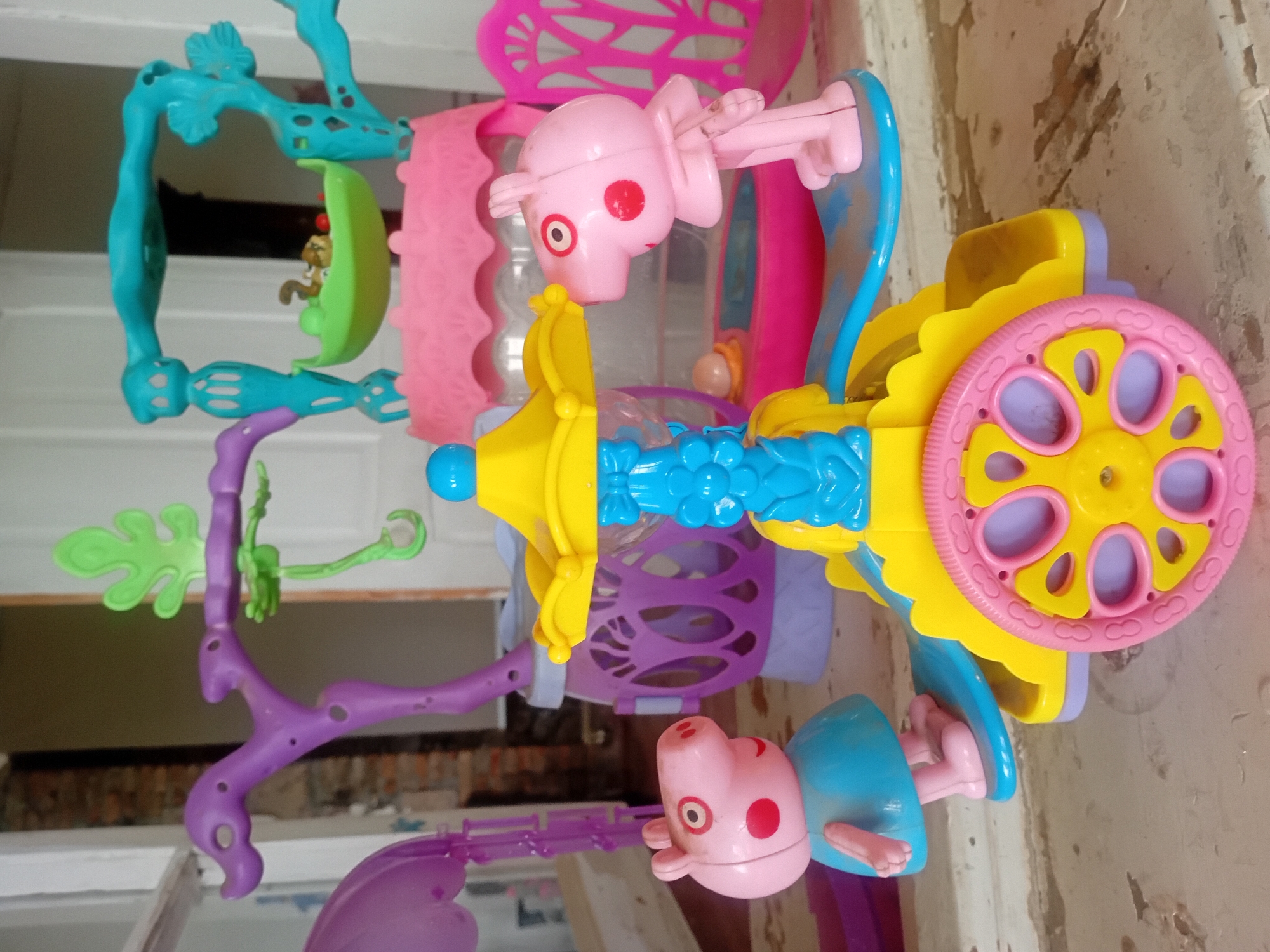This is a medium shot, eye-level, indoor photograph of several plastic toys scattered on a wooden surface. The toys are colorful and appear to be part of a playset. In the foreground, a yellow and blue carousel-like toy is prominent, with a pink wheel and a blue platform. Two pink pig figurines, resembling characters from the show Peppa Pig, are positioned on either side of the carousel. One pig, wearing a blue dress, stands on the left platform. The other pig, also pink, stands on the right platform, facing the carousel.
Behind the carousel, various other colorful plastic structures are visible. To the left, a large purple structure with ornate carvings and a dangling green leaf-like element is present. In the center background, a pink castle-like structure is visible, with a teal frame above it, holding a small green boat with a tiny monkey toy and some red balls inside. To the right, a pink chair with an ornate design is partially visible. The background is out of focus, showing hints of a wall and doorway, suggesting an indoor setting. The wooden surface appears weathered and worn.