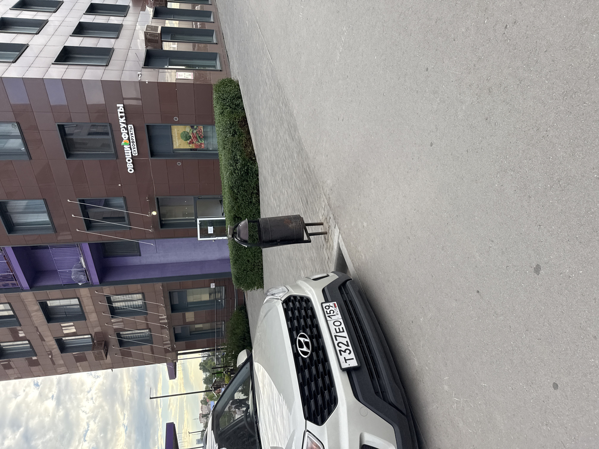 In the city of Perm, Russia, a silver Hyundai Creta is parked on a paved area in front of a multi-story building. The building has a dark brown facade with a purple accent on one section. Above a ground-floor entrance, a sign reads "ОВОЩИ ФРУКТЫ" (Fruits and Vegetables) with smaller text below it. A black trash receptacle stands to the right of the car. The sky is cloudy. The license plate of the car is visible and reads "T327EO 159 RUS".