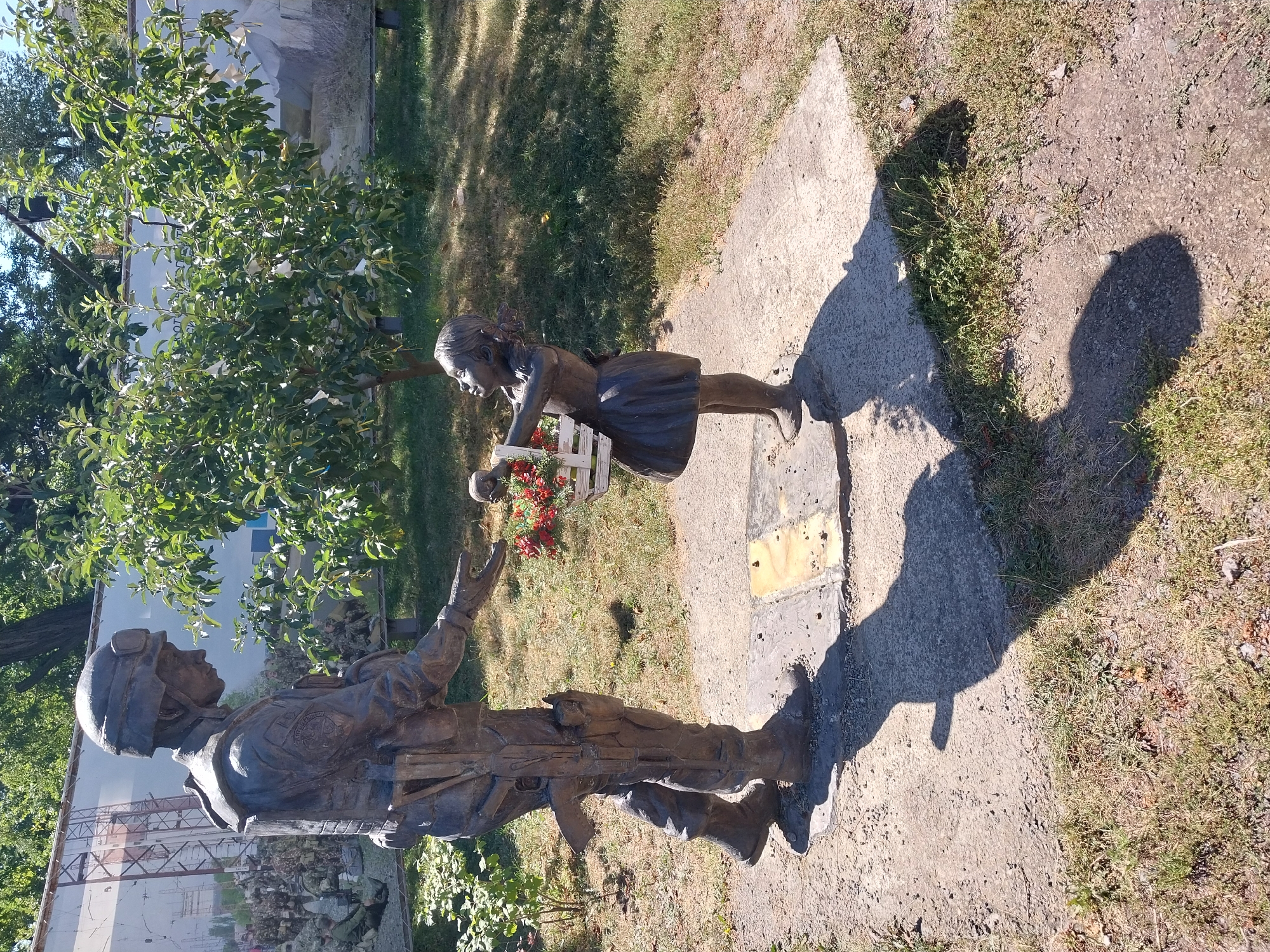 This is a photograph of a bronze statue in the city of Dnipro, Ukraine, depicting a soldier and a child. The soldier, dressed in military gear and helmet, is offering a basket of flowers to a young girl. The girl, wearing a dress, is accepting the flowers with a hopeful expression. The statue is located outdoors in a grassy area with trees and a large banner in the background. The banner appears to be a mural or display related to military themes. The overall scene suggests a somber yet hopeful narrative, possibly commemorating a significant event or reflecting on the impact of conflict on civilians.