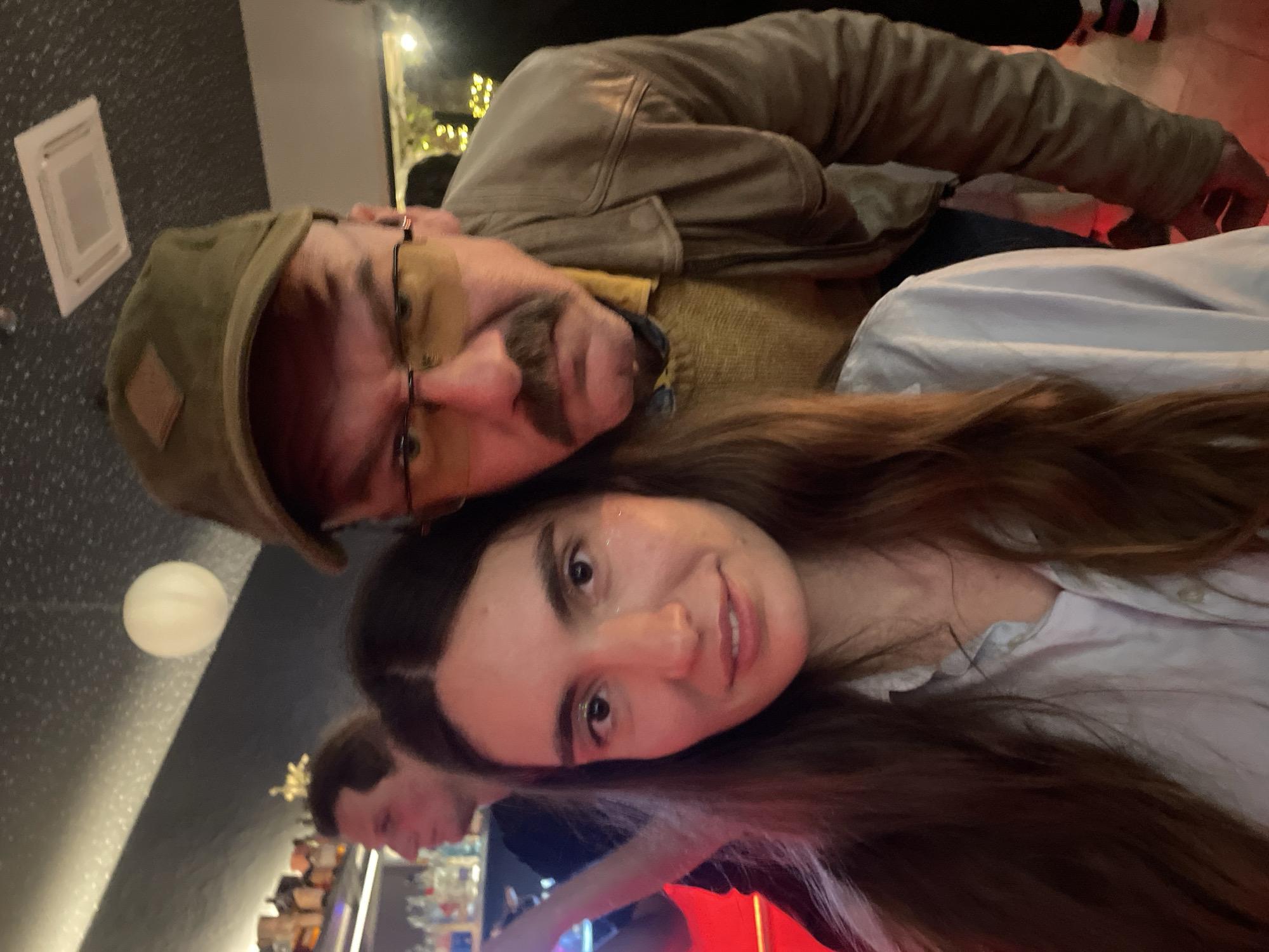 The image depicts a selfie taken indoors, likely in a bar. A woman with long brown hair and a light-colored shirt is in the foreground. Behind and slightly above her is a man wearing a tan jacket, green hat, and glasses with a prominent mustache. He is leaning towards her. In the blurred background, a bar setting is visible, including shelves with bottles, lights, and another person. The location cannot be determined as Boreti, Montenegro, with the context provided.