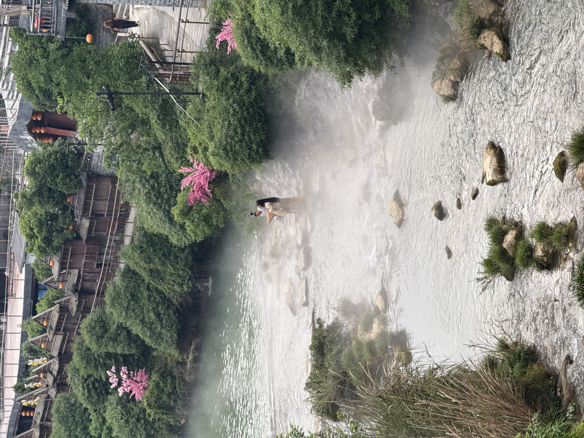 The image presents an elevated view of the Tuojiang River flowing through Furong Ancient Town in Hunan Province, China. Dense foliage, including trees and pink flowering shrubs, lines the riverbanks. Traditional wooden buildings with orange lanterns are visible along the left bank. In the middle of the river, a person is standing and facing the river, wearing a gold-colored robe and black hair. The river water is flowing, with scattered rocks and greenery along the edges. The weather appears misty or humid.