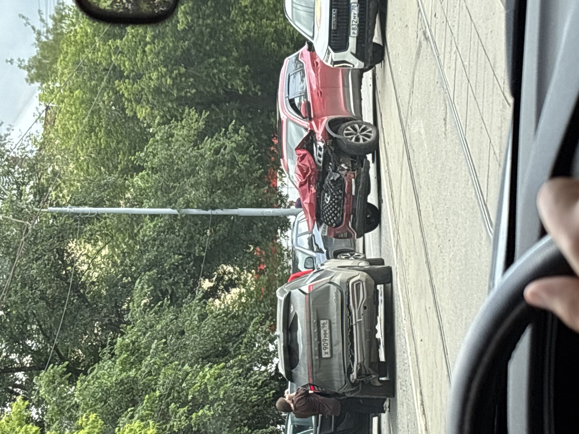 In Yekaterinburg, Russia, a traffic accident has occurred involving at least three vehicles on a city street. A red SUV in the center of the frame has sustained severe front-end damage, with its hood crumpled and airbags deployed. To its left, a dark-colored SUV has damage to its rear bumper and a broken rear window. A person in dark clothing stands next to the dark SUV, looking towards the red SUV. To the right of the red SUV, a white SUV is visible with visible damage to its front grille. The scene is set against a backdrop of green trees and a cloudy sky. The road appears to have tram tracks. The image is taken from inside another vehicle, with a steering wheel and a hand visible in the foreground.