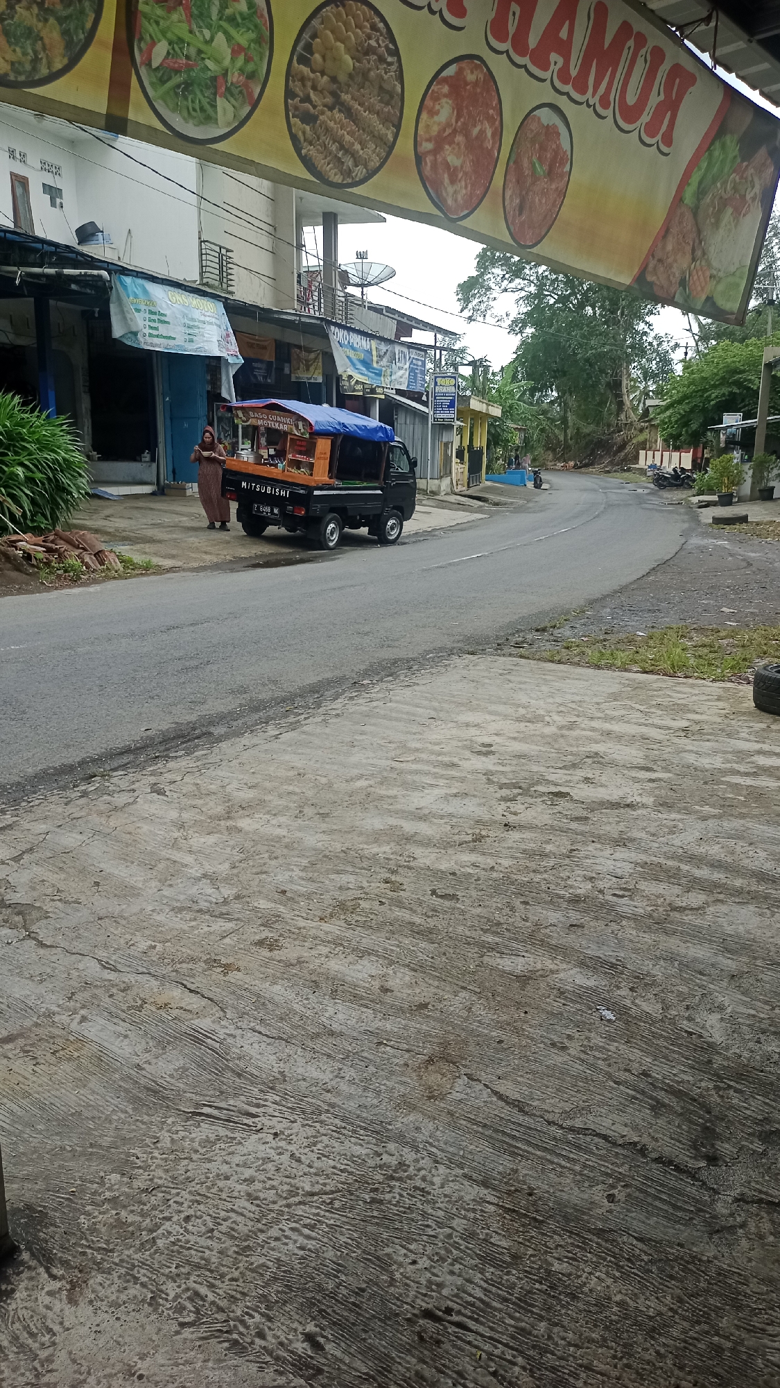 The image shows a street scene in Karyamandala, Indonesia. A small, black Mitsubishi pickup truck, modified into a food cart with an orange and black body and a blue tarp covering, is parked on the side of a paved road. A woman wearing a long brown dress stands near the driver's side of the truck, holding a mobile phone. She appears to be looking at the phone. In the background, several businesses with signs are visible, including one that reads "KOKO RAMA". A large banner with images of food hangs above the storefronts. The road curves to the right and leads uphill. Trees and vegetation line the sides of the road, and several motorcycles are parked in the distance. The foreground consists of a textured concrete surface.